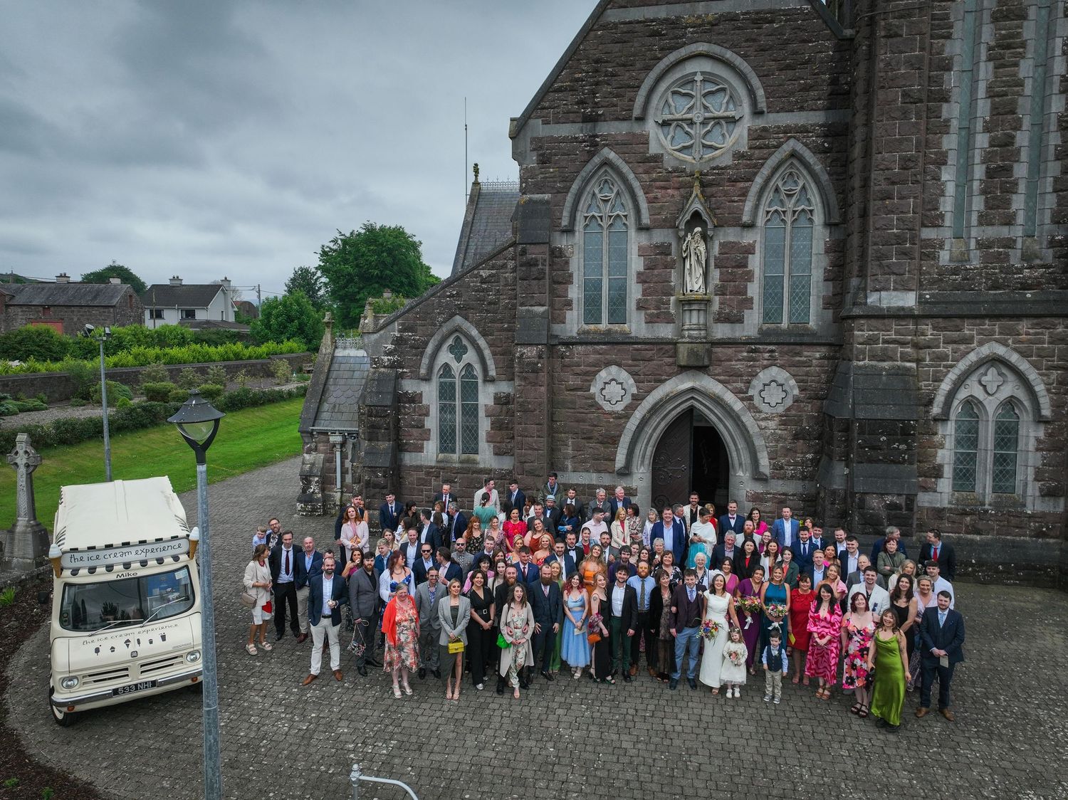 Large wedding group photo outside a historic stone church with a vintage white van on a cloudy day.