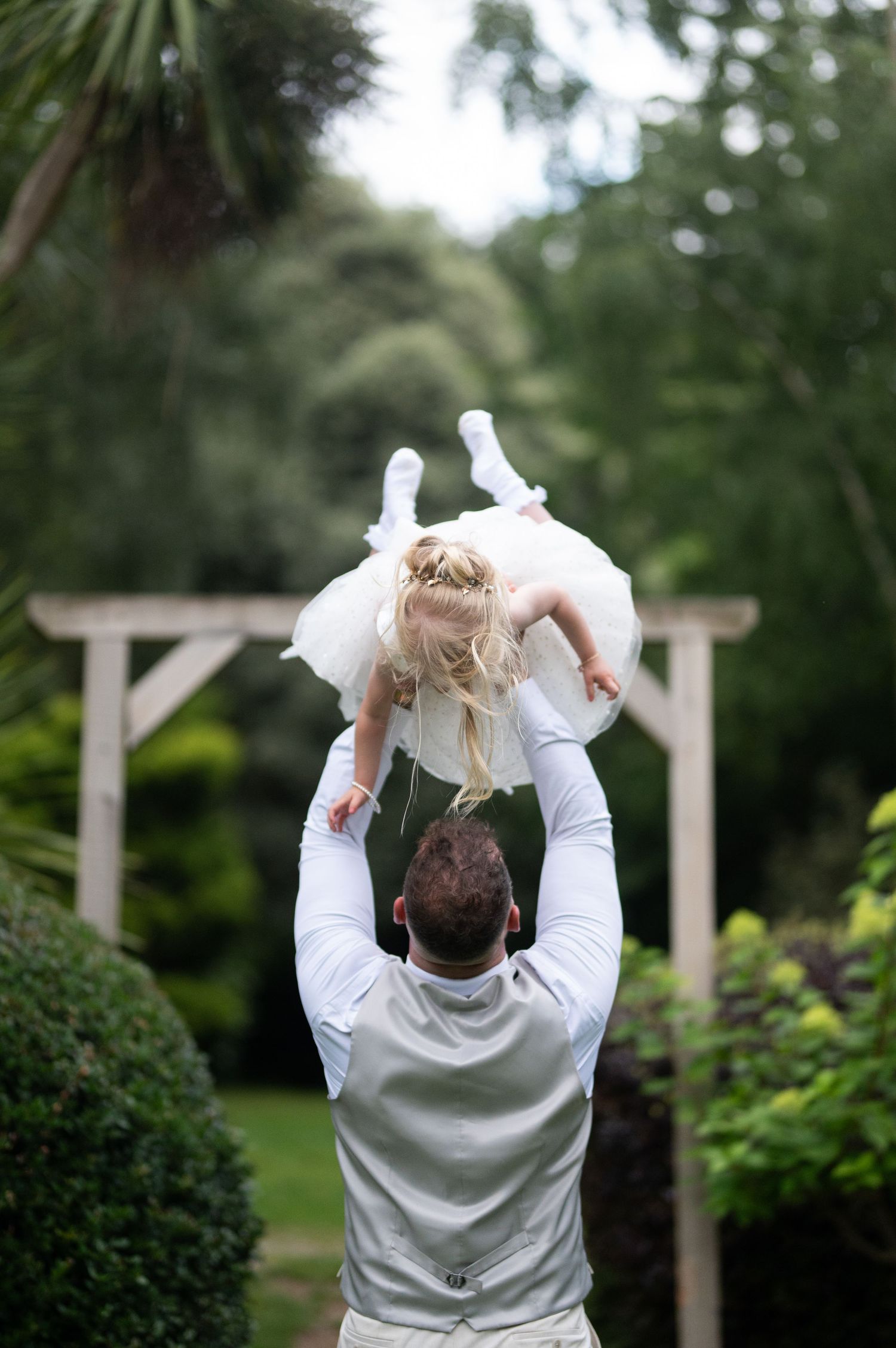 Groom in grey vest lifting young flower girl in white dress overhead in lush garden setting.