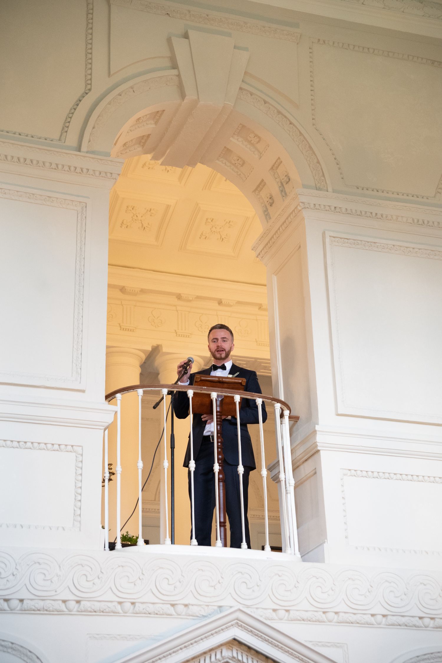 Man in navy suit speaking at a podium on an elegant white balcony inside a grand classical building.