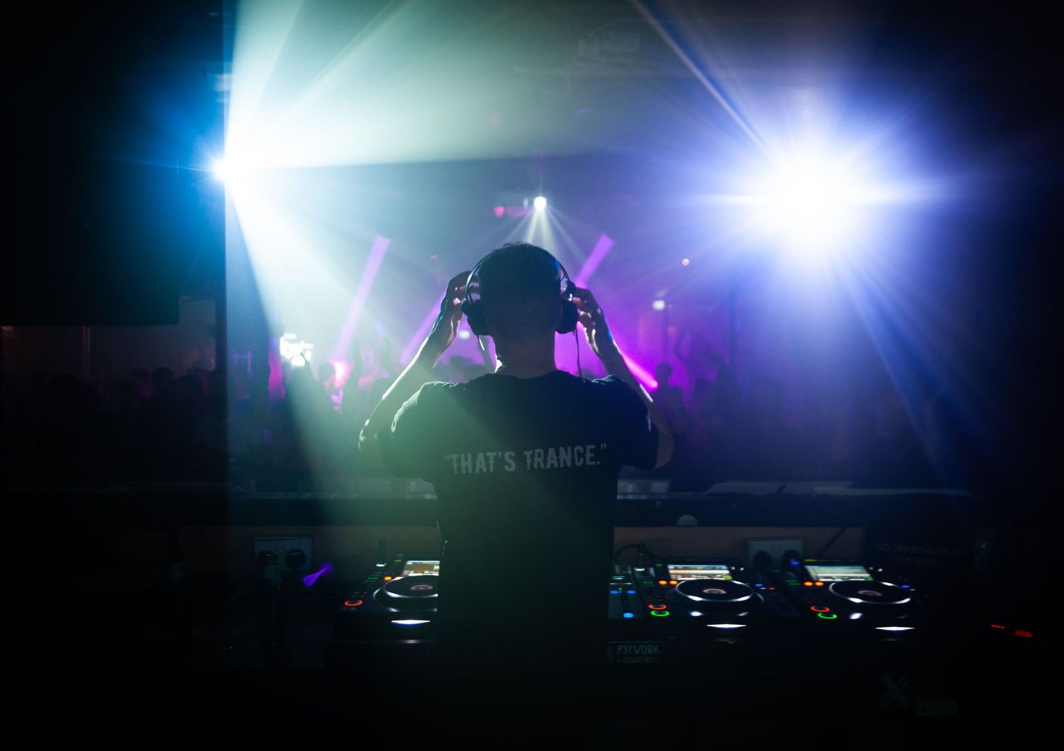 Silhouetted DJ with headphones performing at trance music event with bright stage lights.
