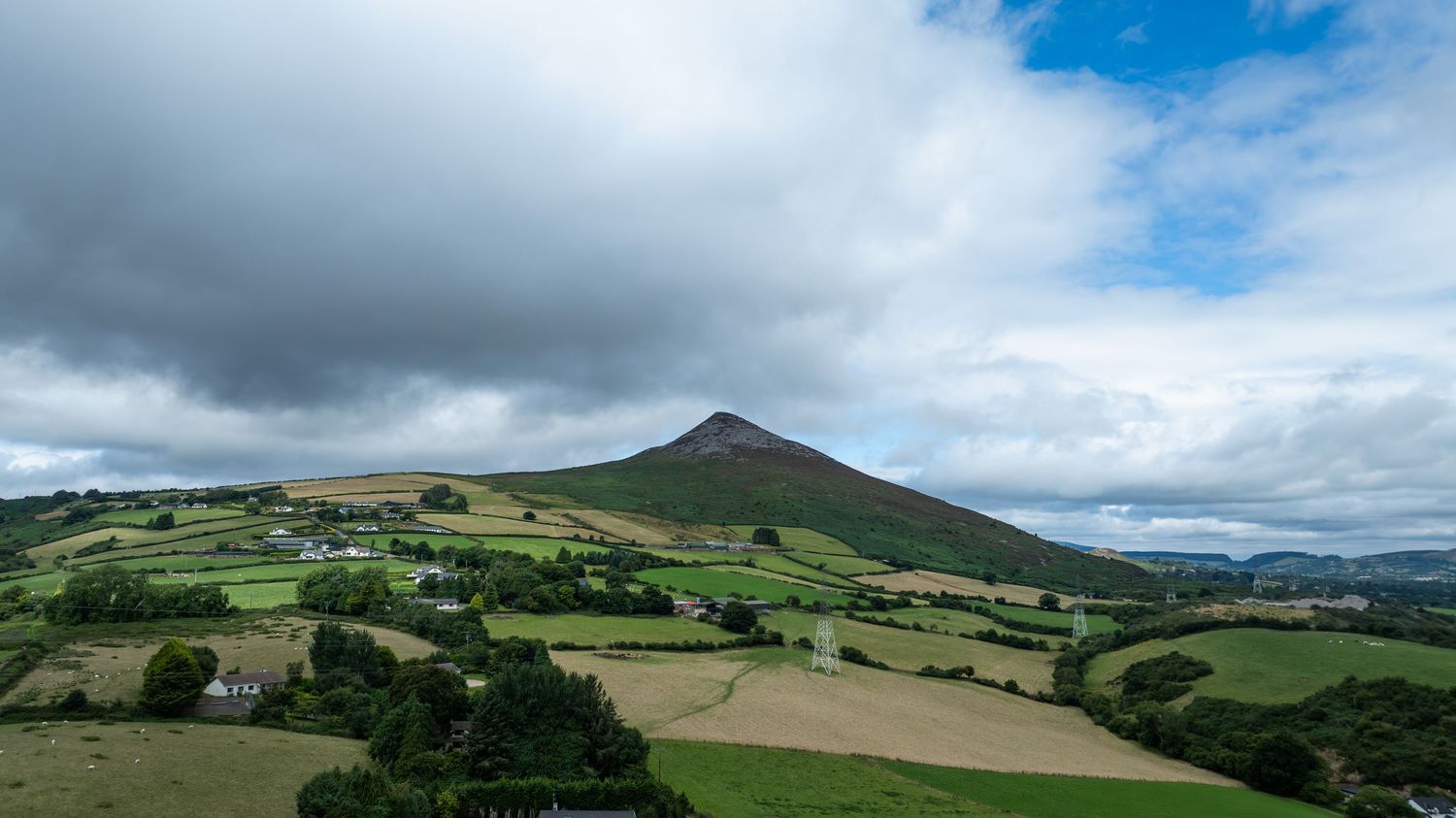 Aerial view of a conical mountain peak with green fields and dramatic cloudy sky in Ireland.