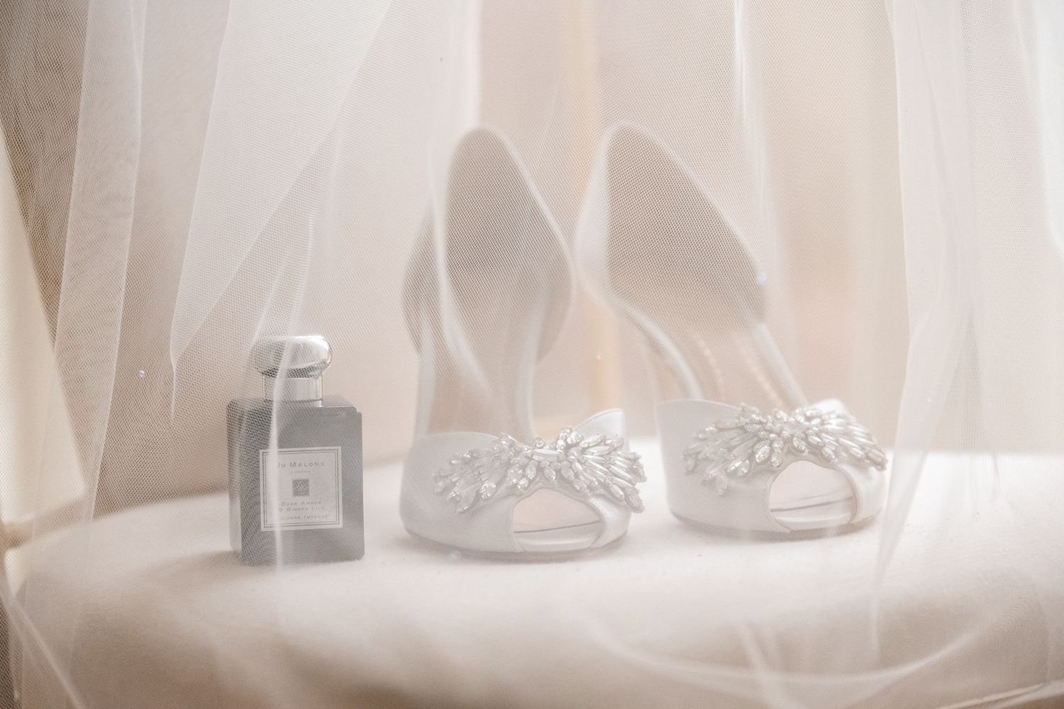 White bridal heels adorned with crystal embellishments beside a Jo Malone perfume bottle draped with a sheer wedding veil.