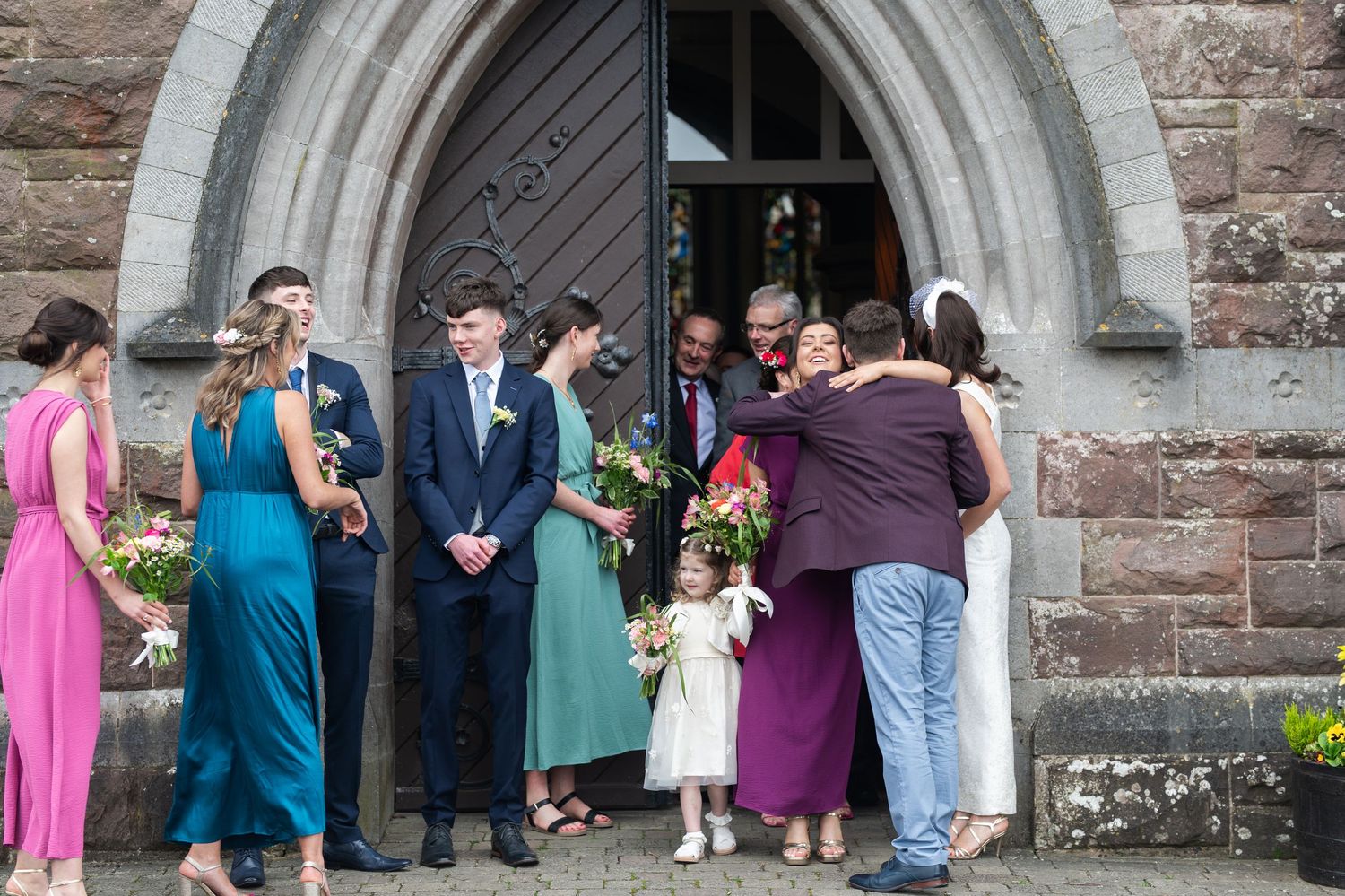 Wedding party gathered outside a stone church, bride and groom with bridesmaids in teal and pink dresses receiving congratulations.