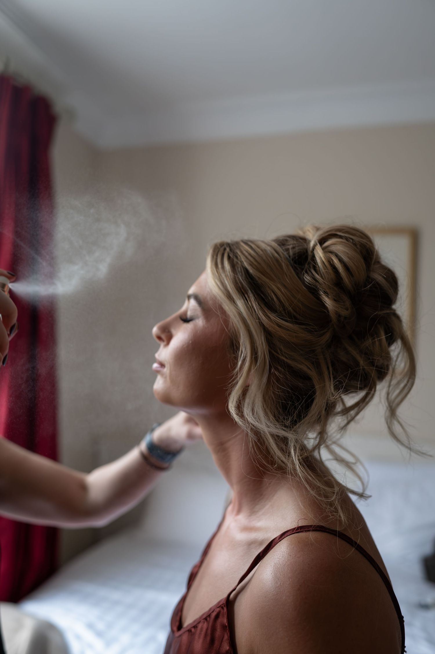 Bride getting makeup set with setting spray, elegant updo hairstyle, wedding morning prep.