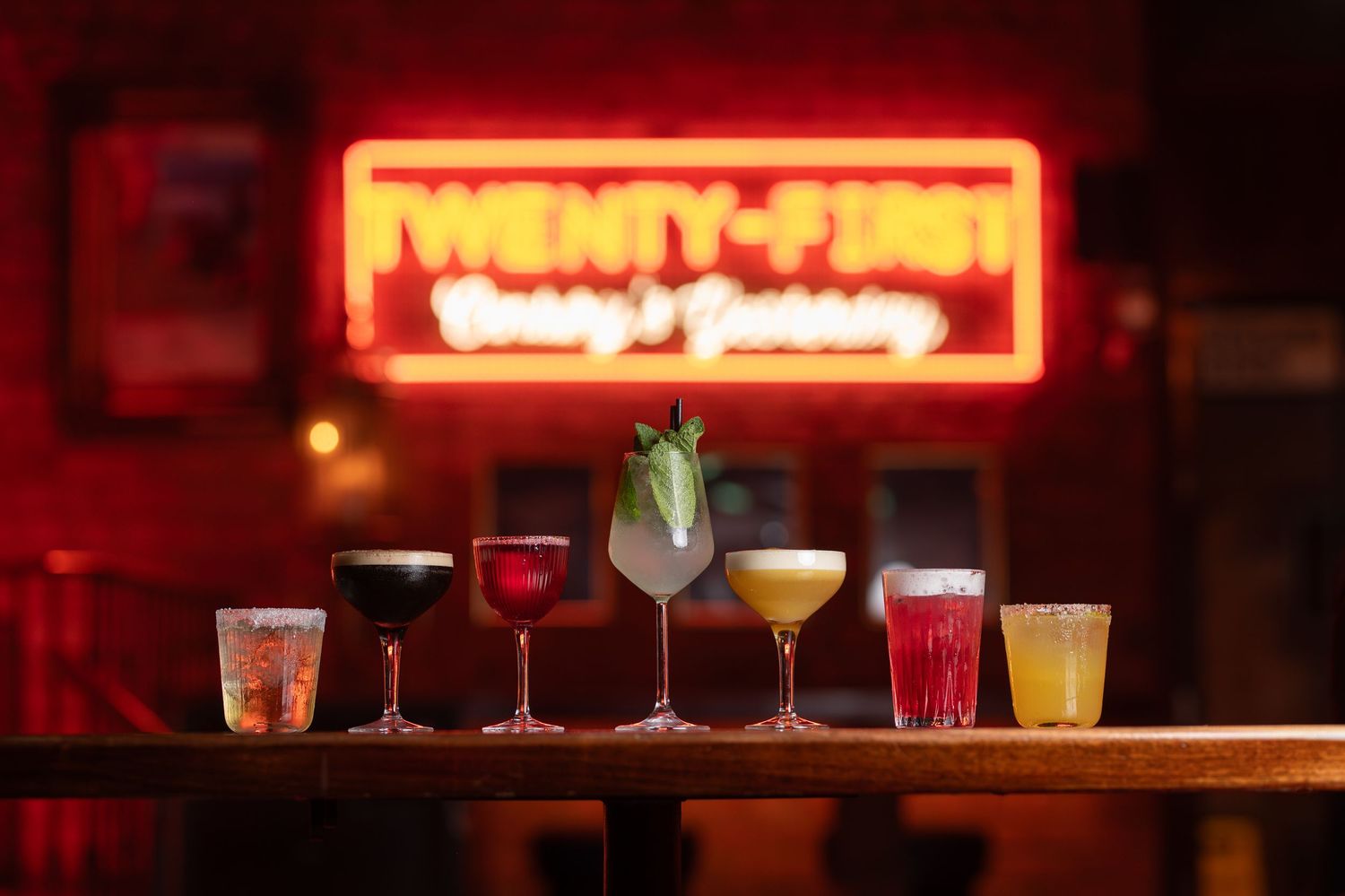Six colorful cocktails displayed on a bar counter beneath a glowing Twenty-First neon sign.