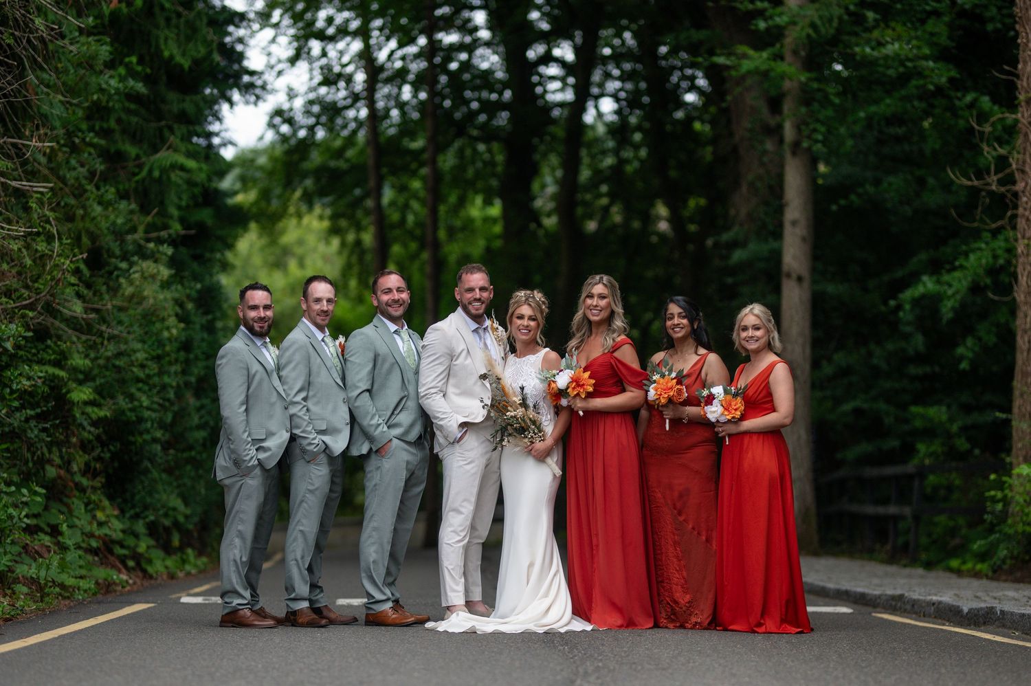 Wedding party posing on road surrounded by trees, bride in white, bridesmaids in red gowns.