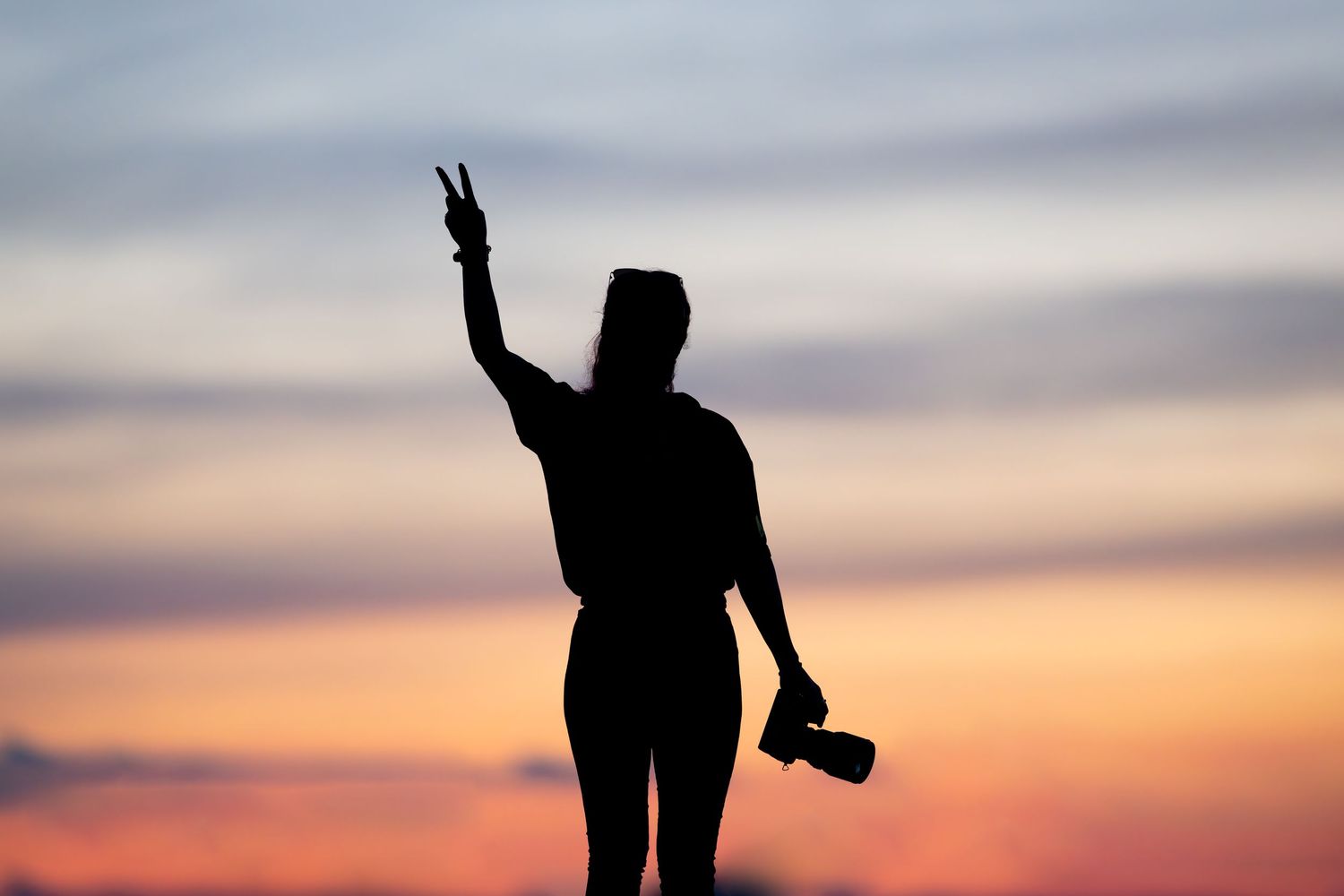 Silhouette of a woman holding a camera and making a peace sign against a vibrant sunset sky.