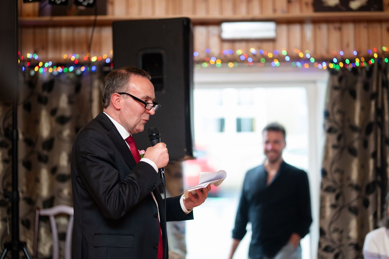 Man in a suit speaking into a microphone at an indoor wedding reception.