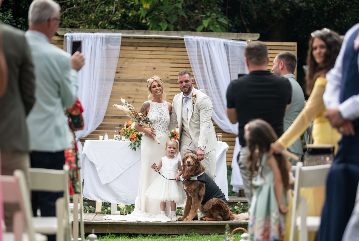 Bride and groom pose with flower girl and dog at outdoor wedding ceremony with white draped wooden arch backdrop.