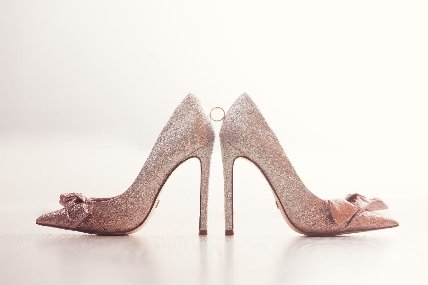 Elegant glittery rose gold bridal heels with bow detail and a wedding ring displayed on white surface.