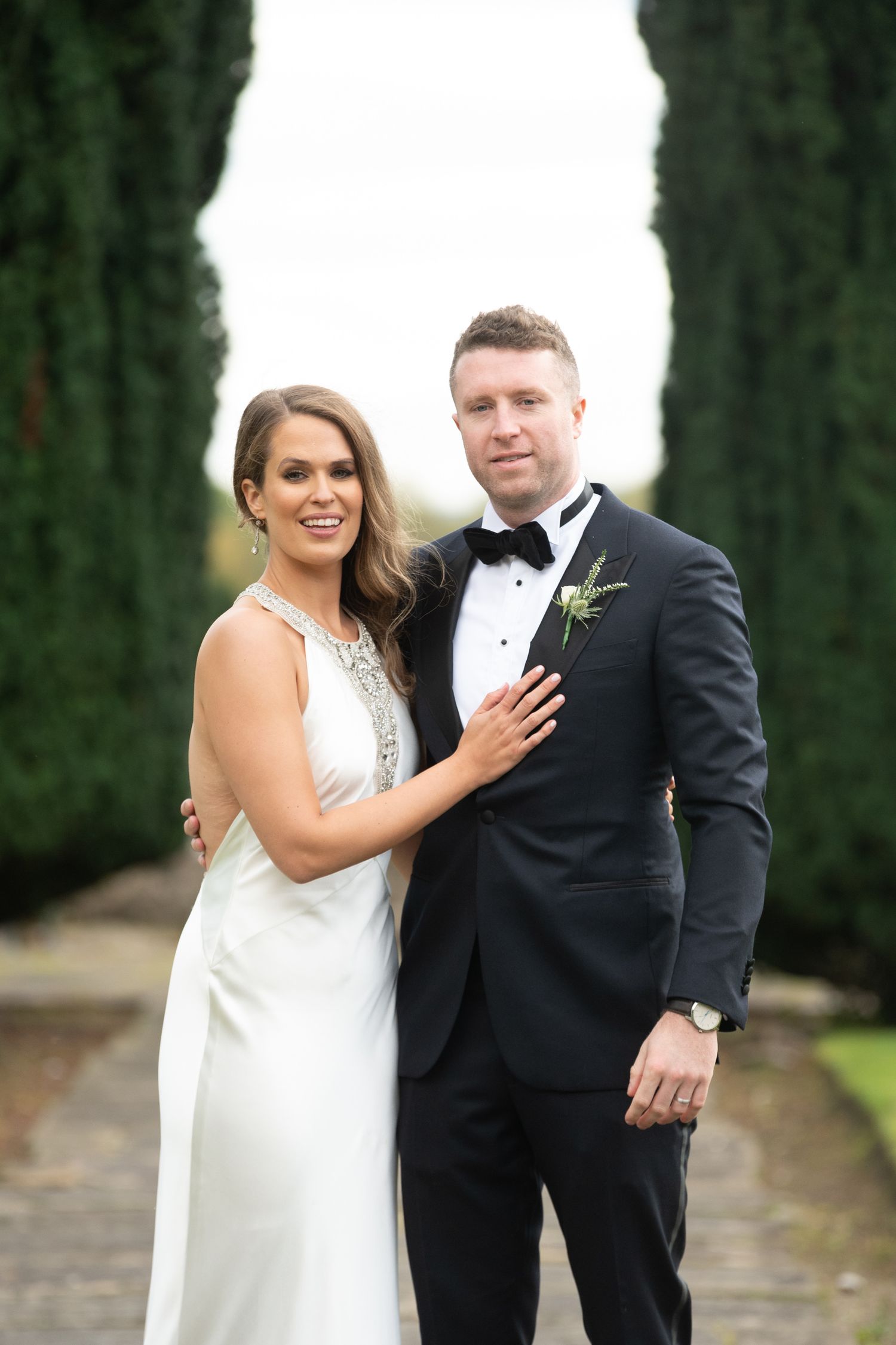 Bride in white gown and groom in black tuxedo pose together outdoors with tall cypress trees behind them.