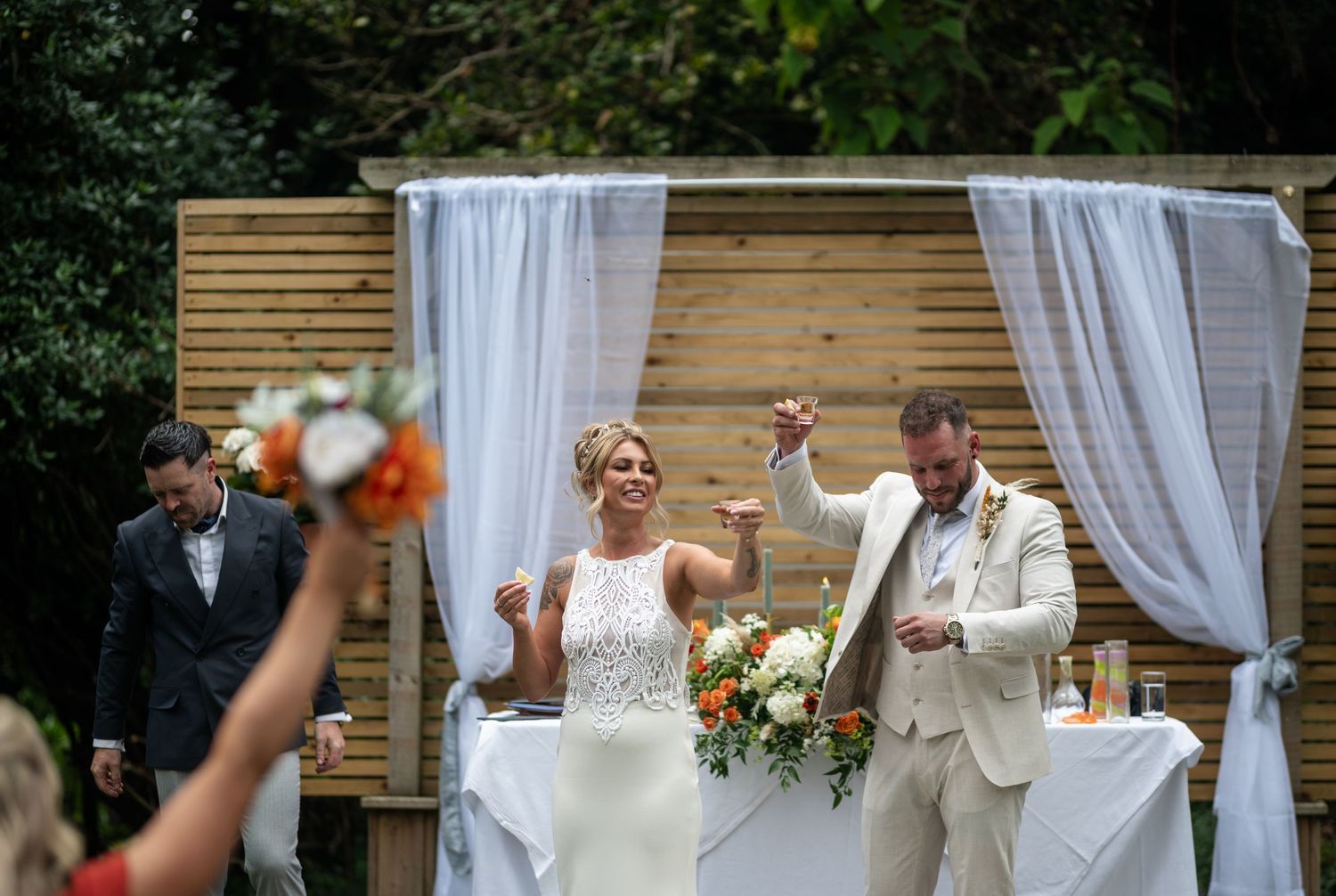 Joyful bride and groom raise hands in celebration at outdoor wedding ceremony beneath white draped altar.