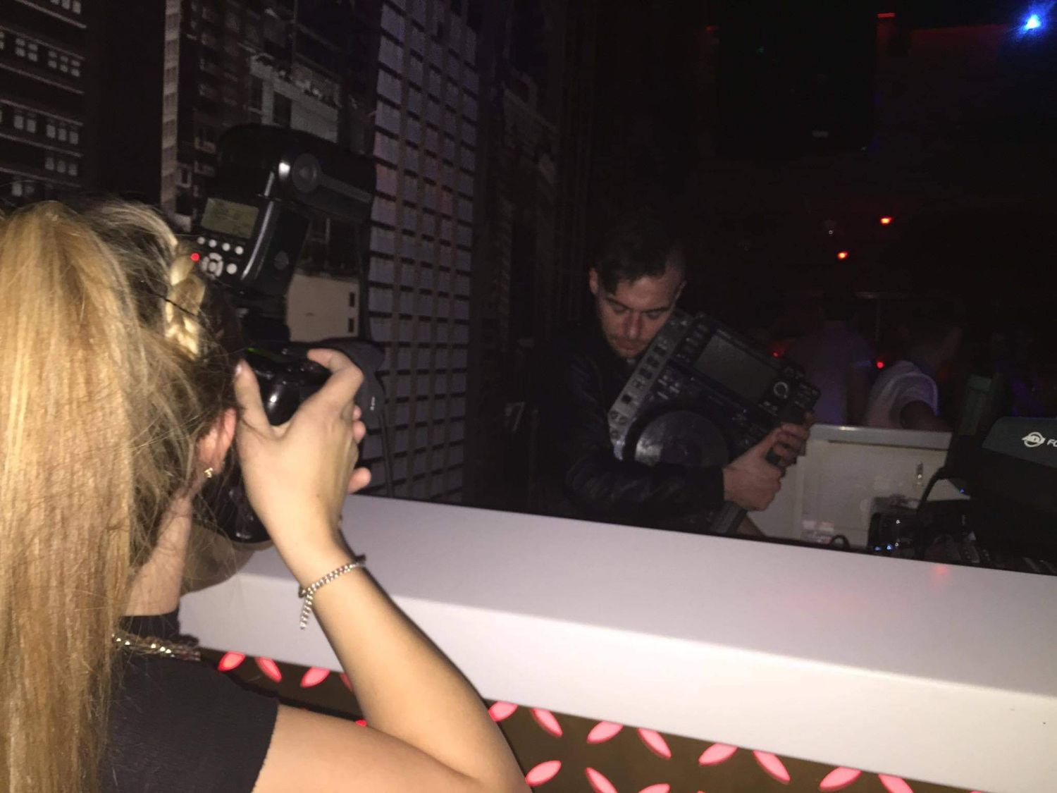 A blonde woman photographs a man with a camera at a dimly lit nightclub bar.