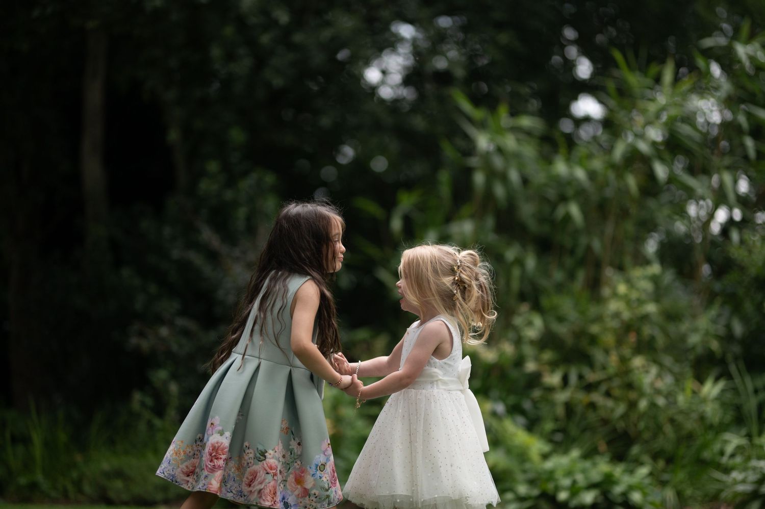 Two young flower girls in floral and white dresses holding hands in a lush garden setting.