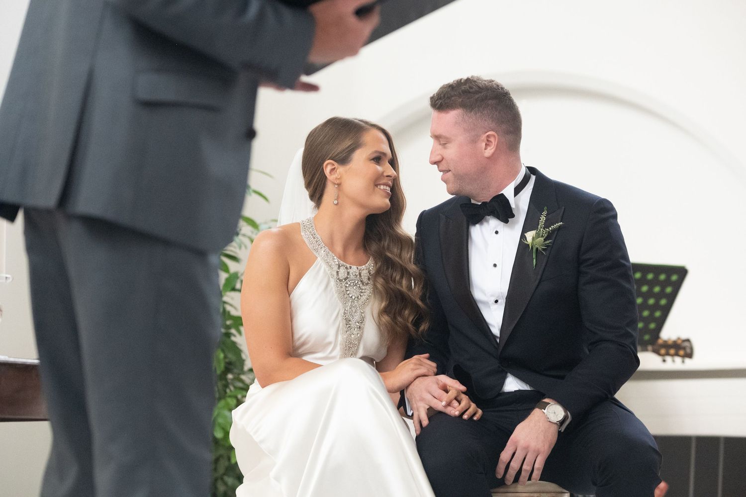 Bride and groom smiling at each other during wedding ceremony, groom in black tuxedo, bride in white gown.