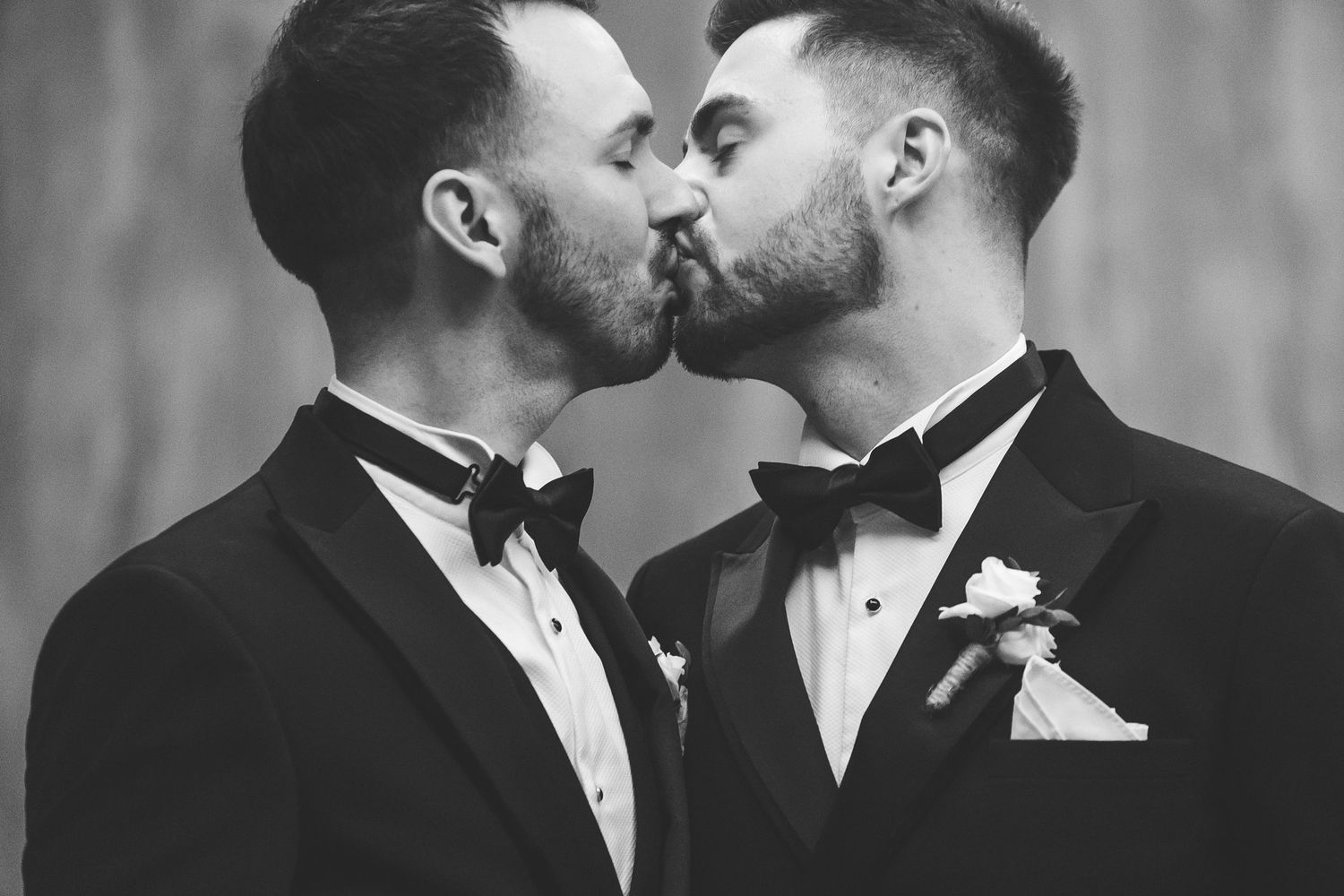 Two grooms in black tuxedos sharing a kiss on their wedding day in a romantic black and white photo.