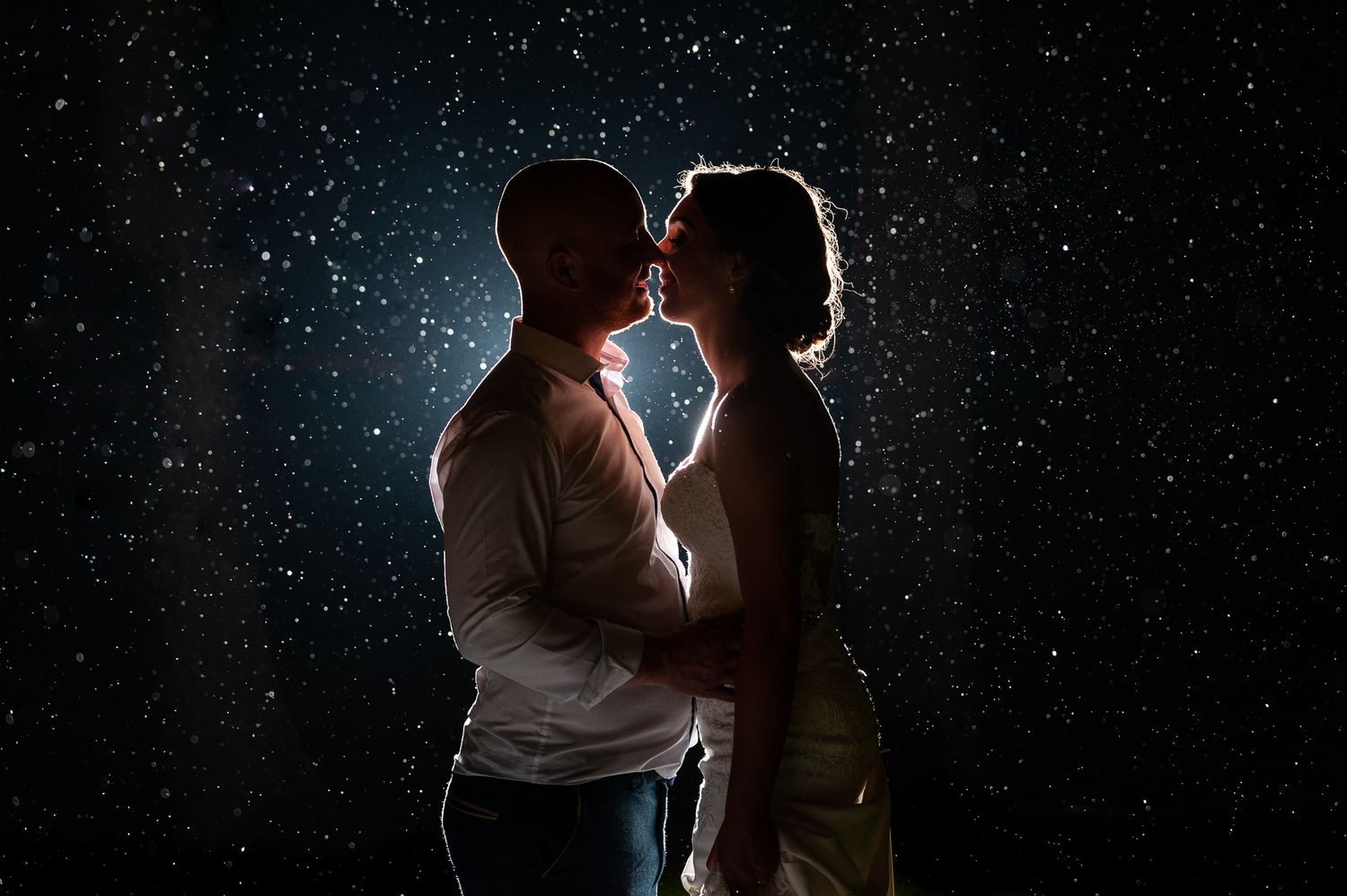 Romantic couple sharing a kiss in the rain illuminated by dramatic backlight against dark night sky.