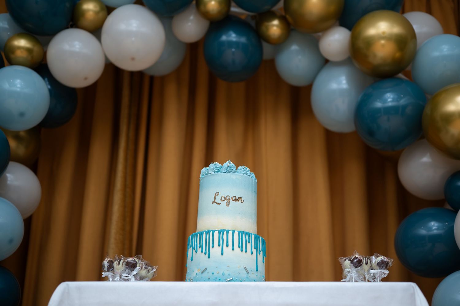 Blue and white two-tier cake reading Logan under a balloon arch decoration.
