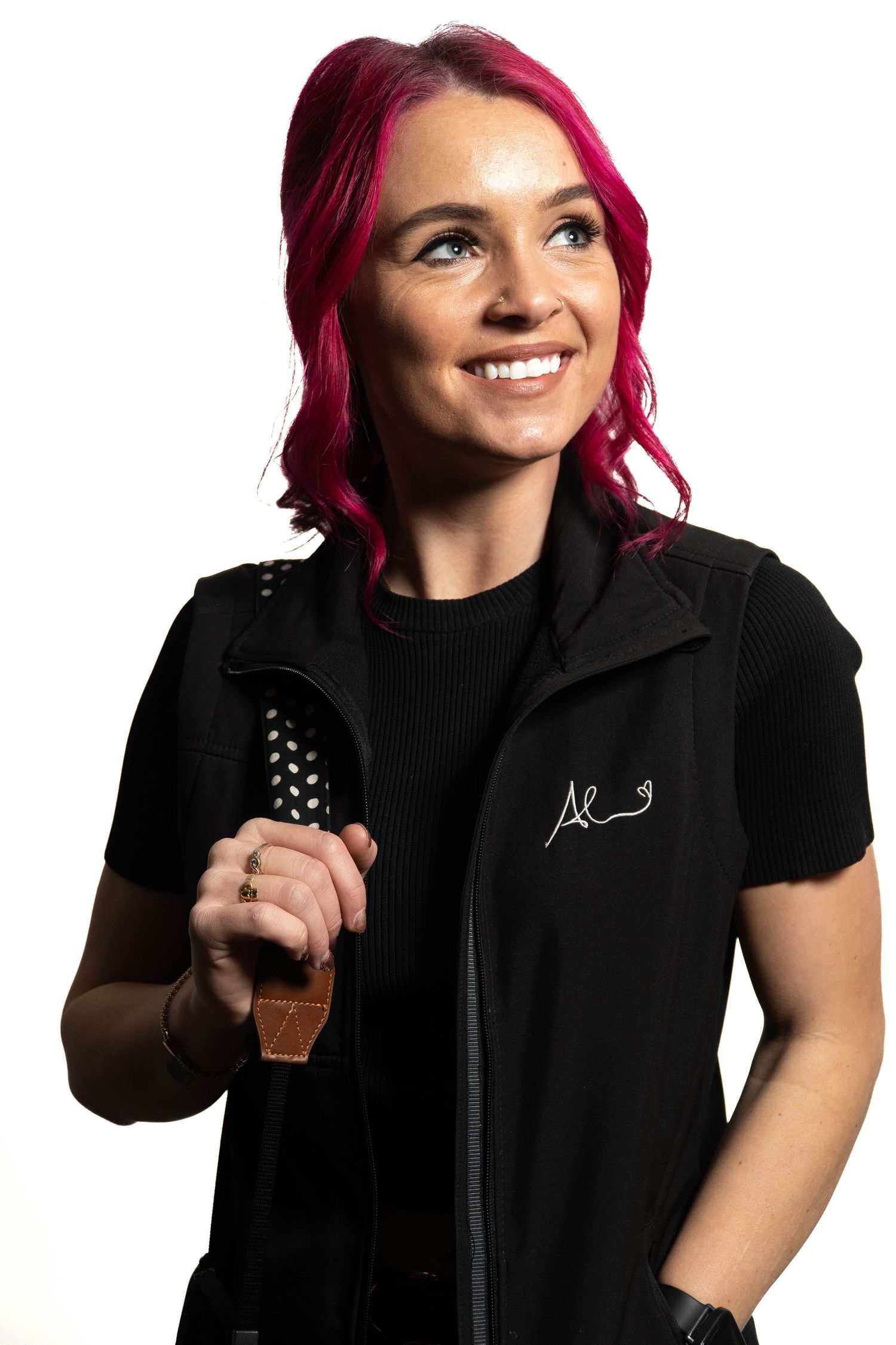 Smiling woman with bright pink hair holding grooming brush wearing black vest on white background.