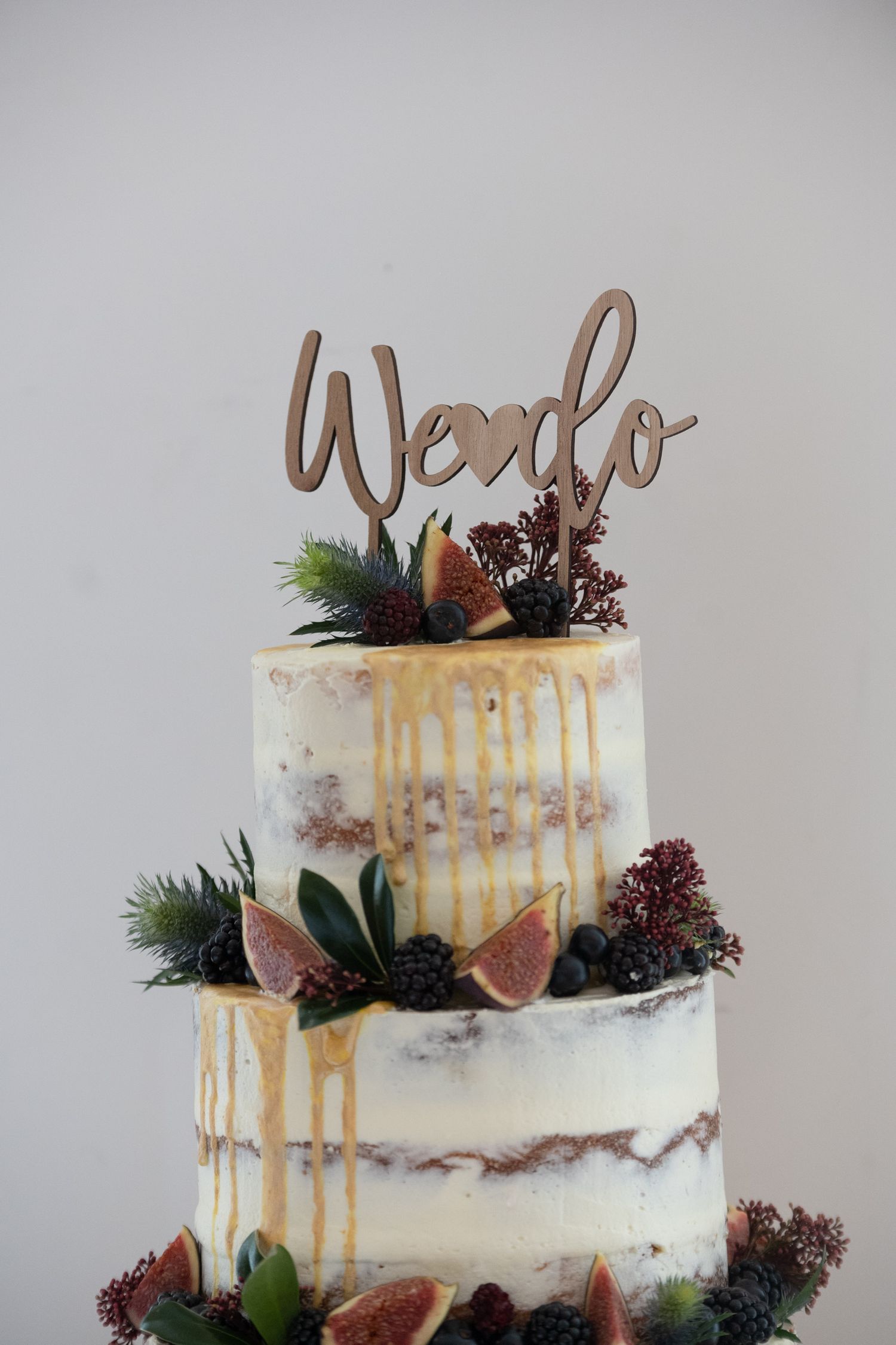Two-tier naked wedding cake with caramel drip, fresh figs, berries, greenery, and a wooden We Do cake topper.
