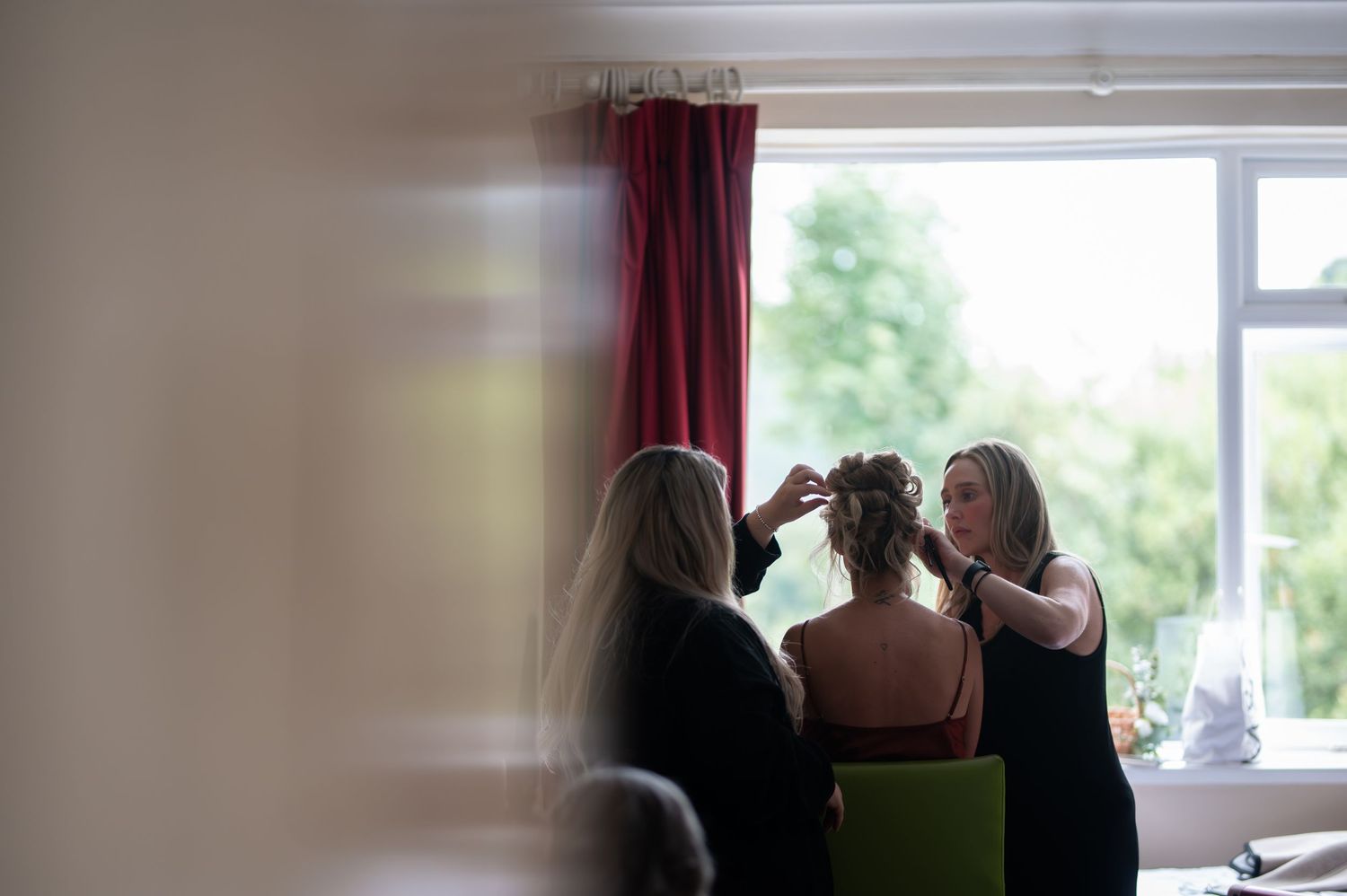 Two women styling a bride's hair near a window during wedding preparations.