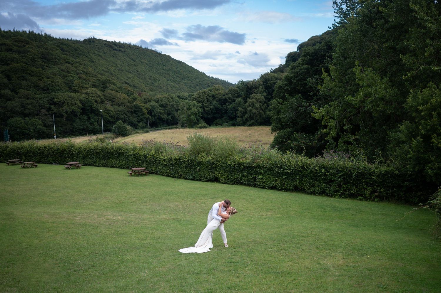 Bride and groom share a romantic embrace on a green lawn with scenic wooded hills behind them.
