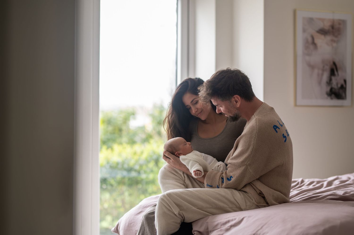 Couple sitting on bed by window tenderly holding their newborn baby at home.