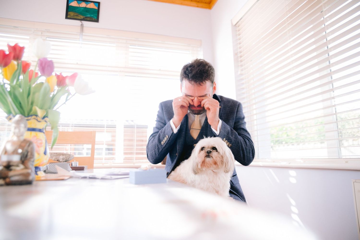 Emotional groom wiping tears at table with small white dog beside him before wedding.