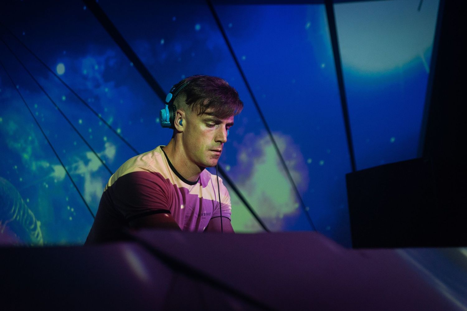 Young male DJ focused on mixing equipment against vibrant blue and green visual backdrop.