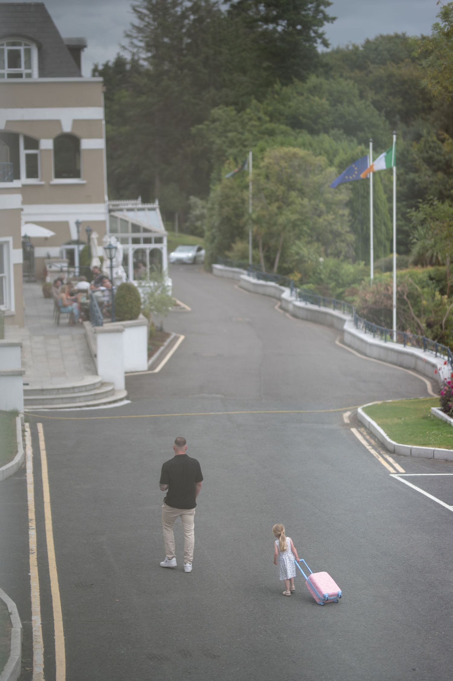 Adult and child walking down a hotel driveway with pink luggage, flags and lush trees in background.