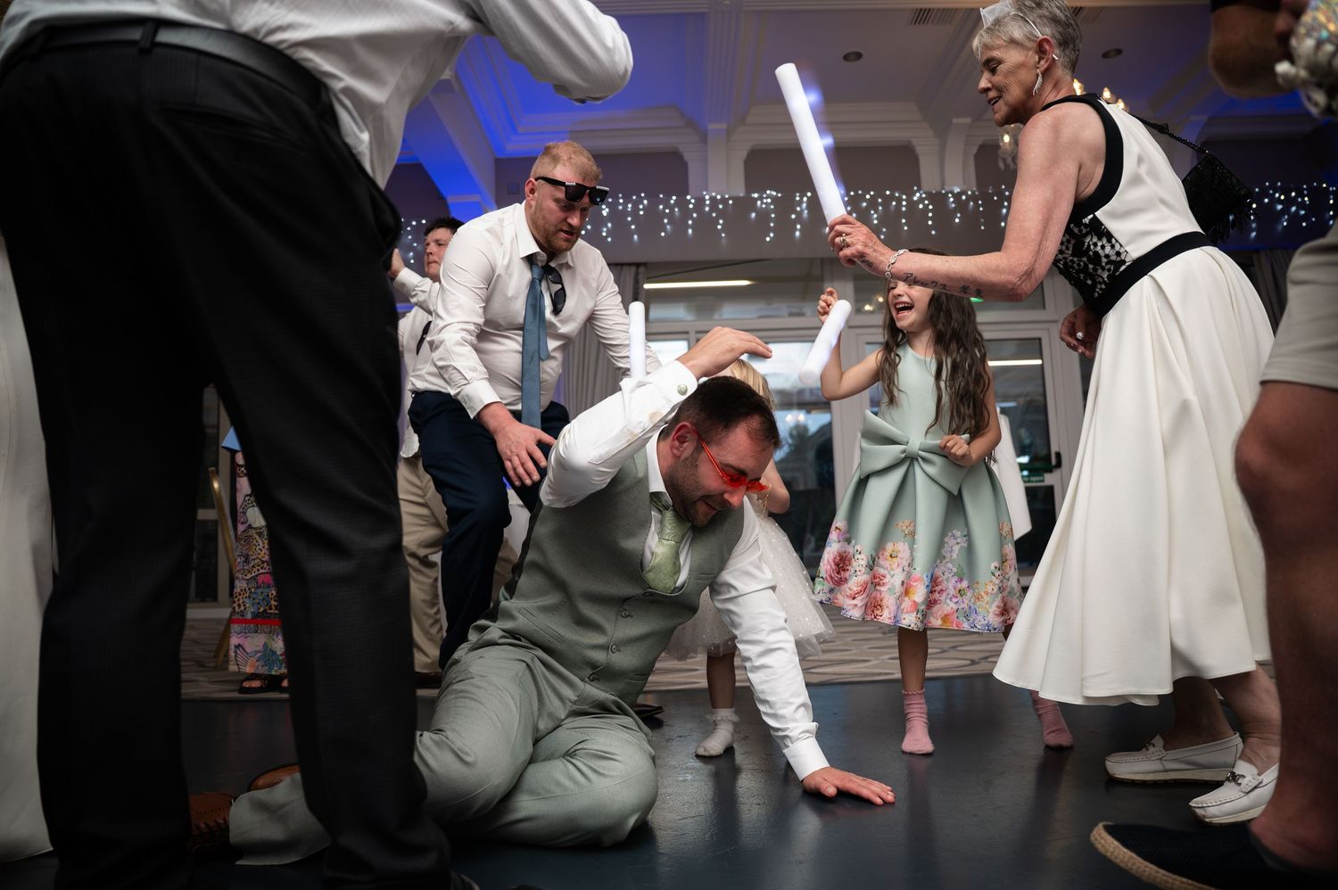 Energetic wedding reception dance floor with groom dancing low surrounded by celebrating guests.