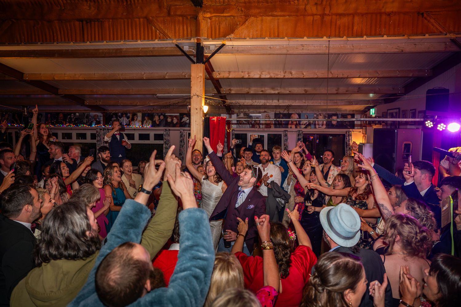 Lively wedding reception dance floor packed with celebrating guests raising their hands in a rustic venue.
