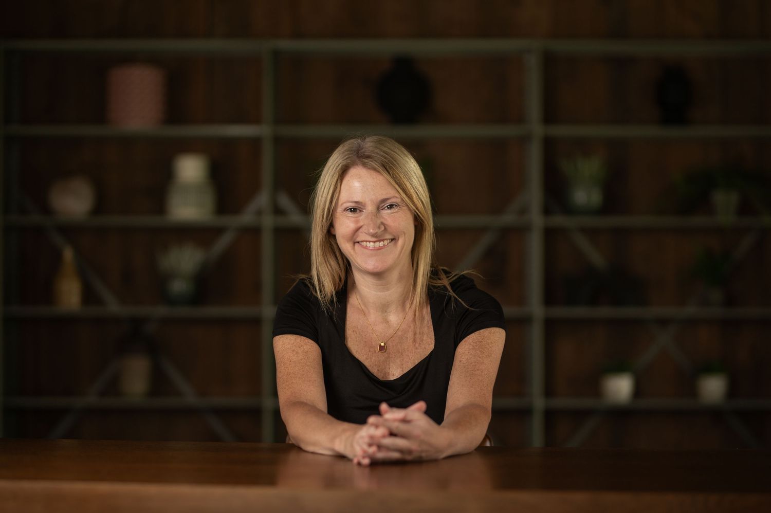 Smiling blonde woman in black top seated at wooden table in modern interior.