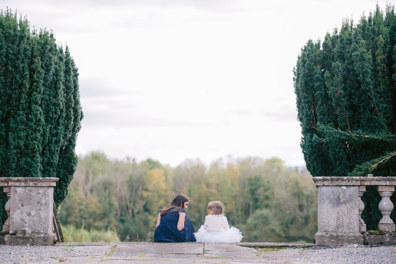 Woman and flower girl sitting together between tall hedges in elegant garden landscape.