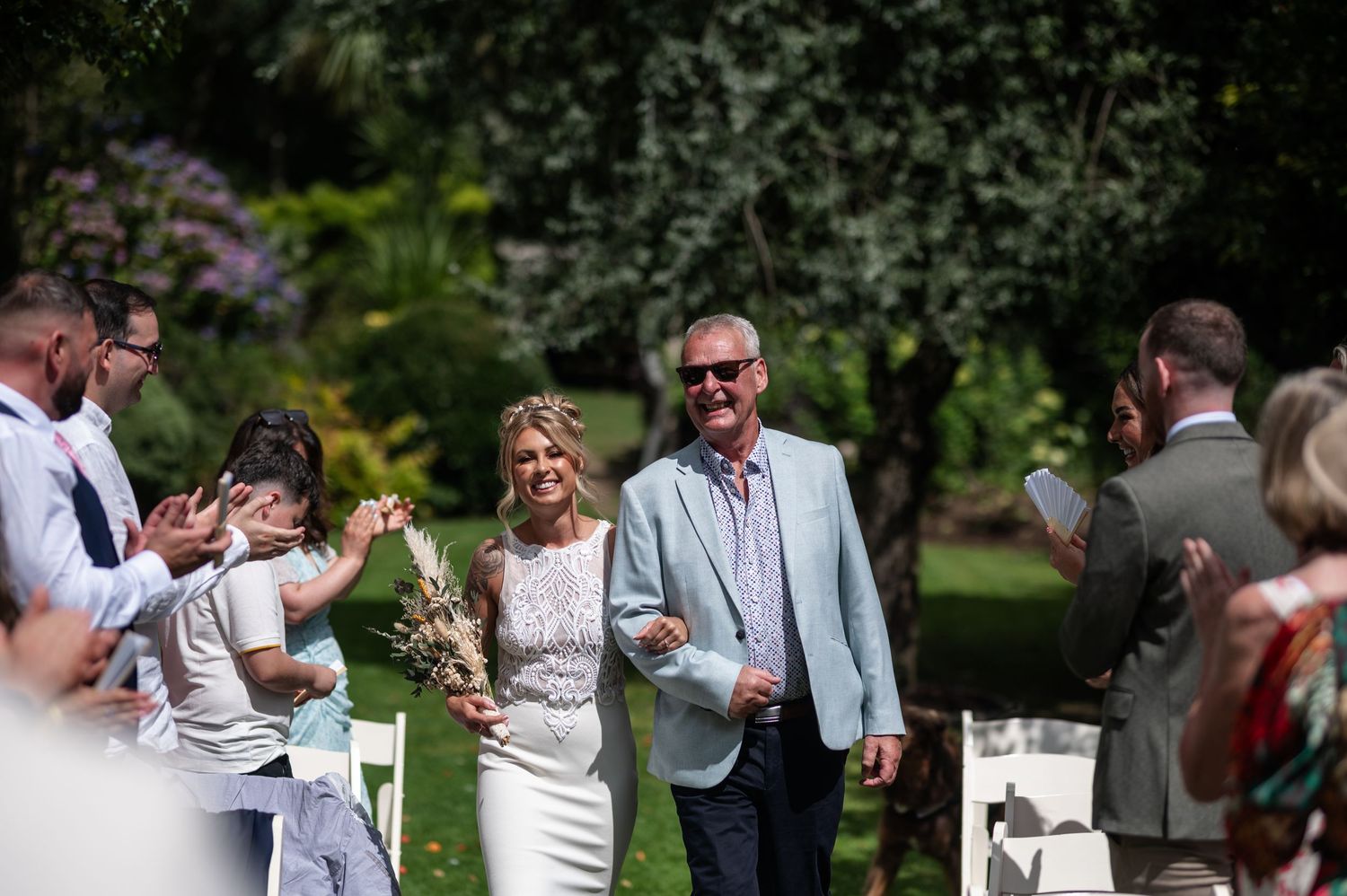 Bride walks down outdoor aisle with escort past applauding guests in sunny garden wedding ceremony.