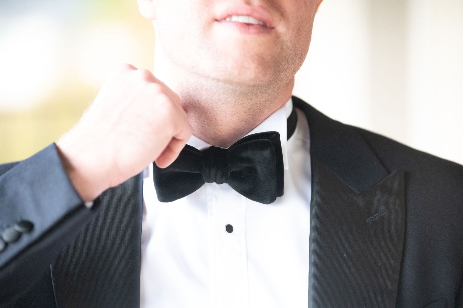 Groom in black tuxedo adjusting his black velvet bow tie before wedding ceremony.