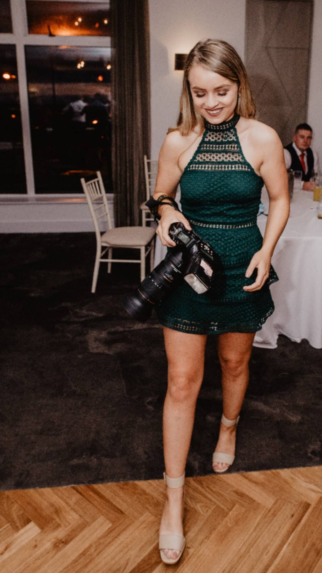 Young woman in green lace dress holding professional camera at elegant indoor event venue.