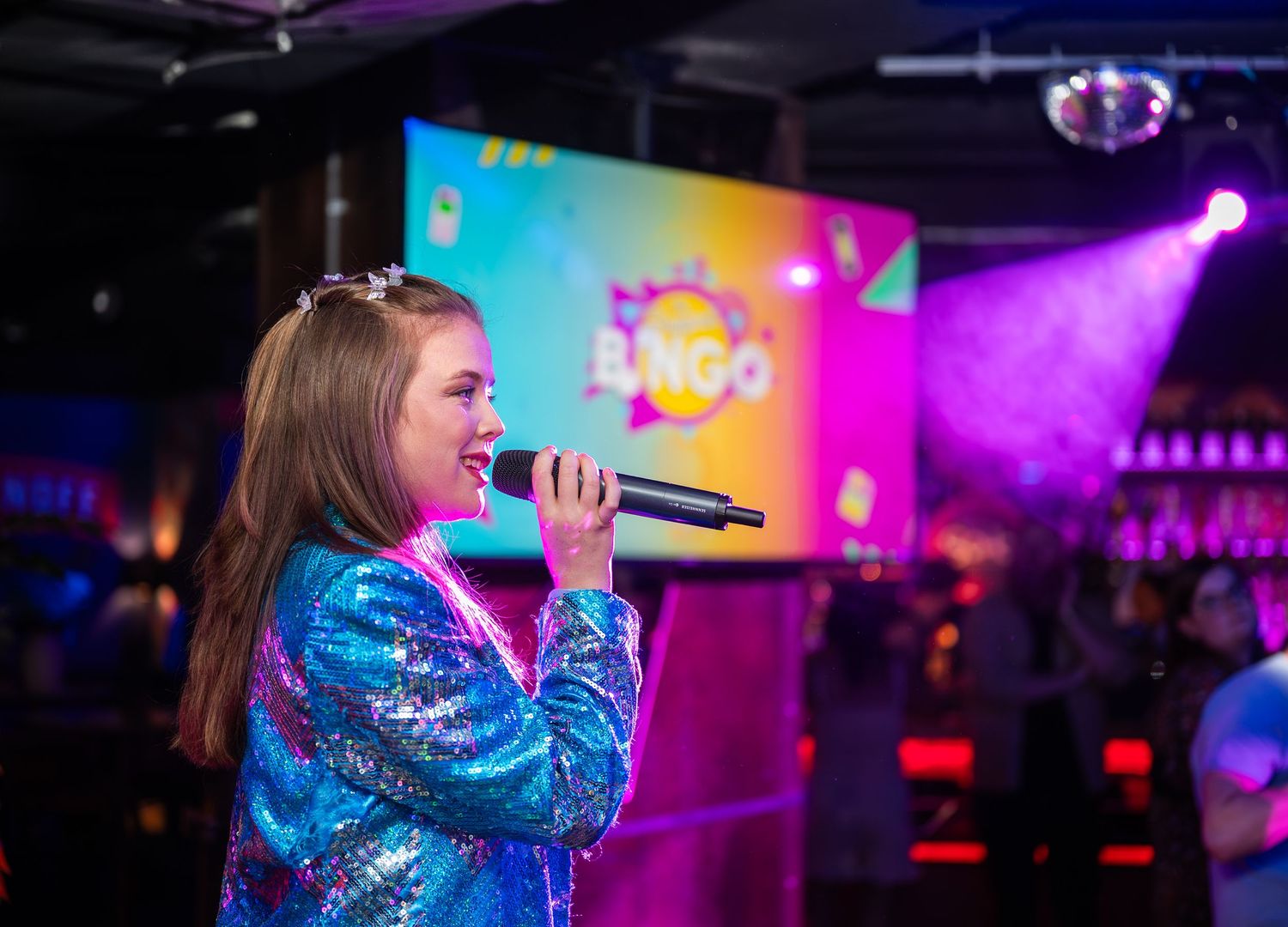 A young girl in a blue sequin jacket singing into a microphone at a bingo event.