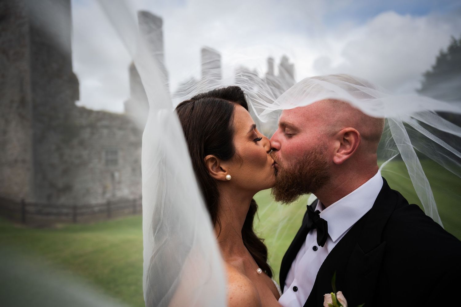 Bride and groom share a romantic kiss outdoors near a historic castle, with the bride's veil blowing in the wind.