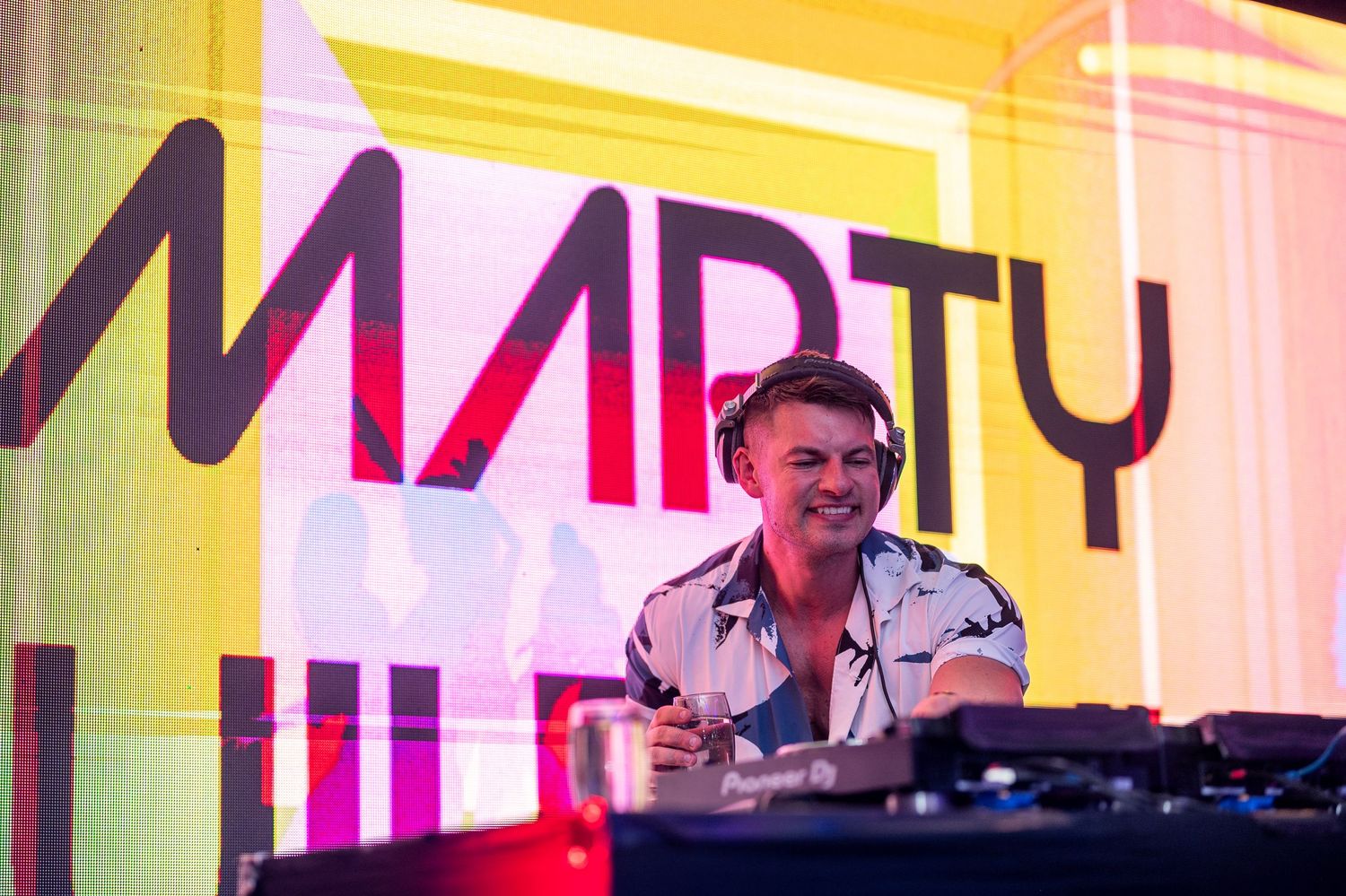 DJ Marty smiling behind Pioneer DJ decks performing live on stage with colorful yellow and pink branded backdrop.