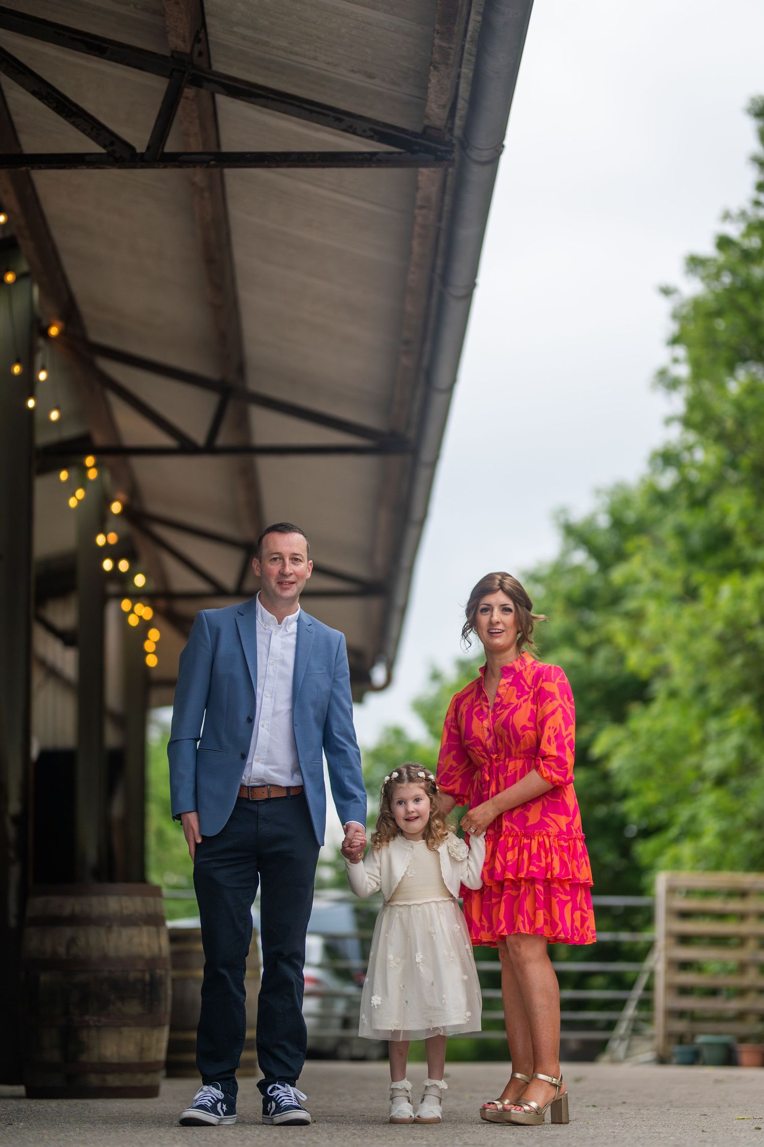 Family of three posing together outside a rustic venue with string lights.