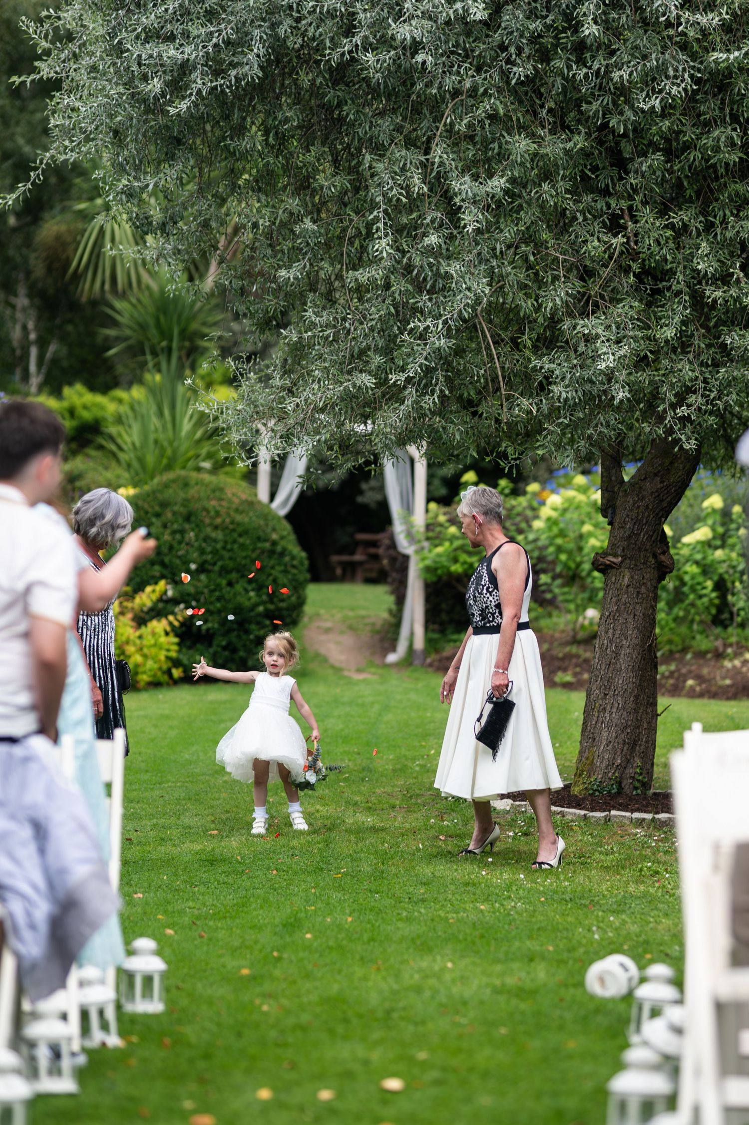 A young flower girl in a white dress plays on a lush green lawn during an outdoor wedding ceremony.