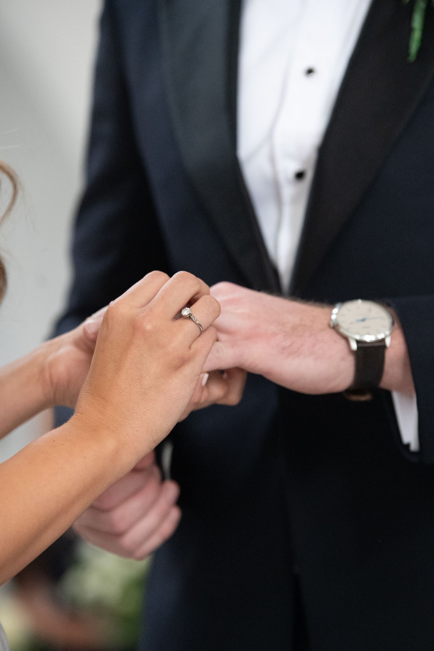 Bride placing a diamond engagement ring on groom's finger during wedding ceremony, groom wearing black tuxedo and watch.