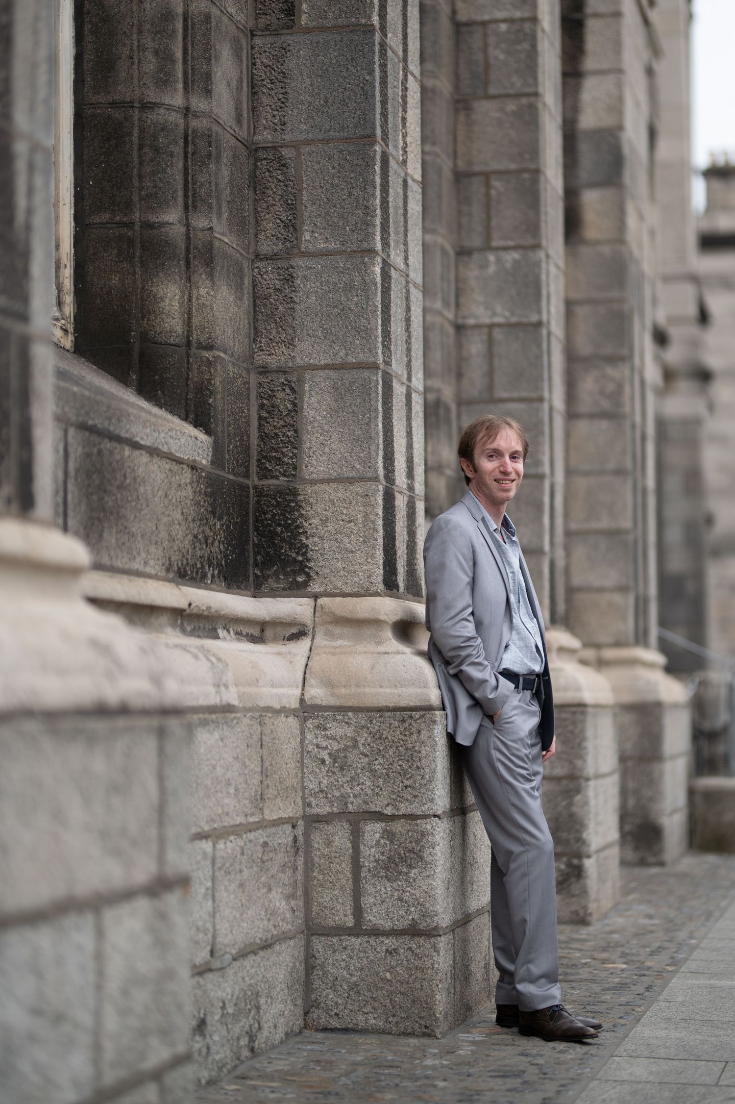 Man in gray suit leaning against stone building wall, smiling confidently in a classic architectural setting.