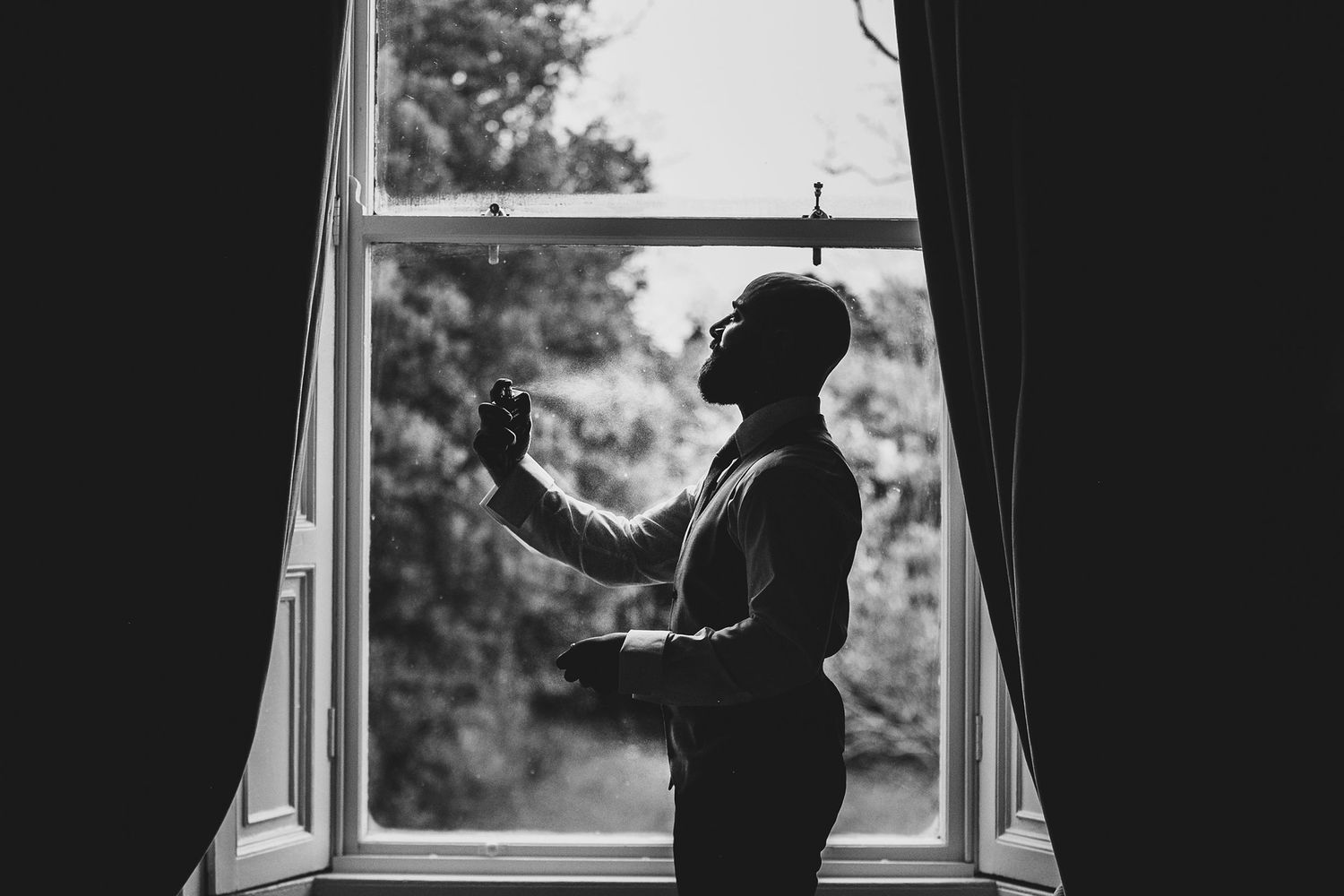 Groom silhouetted by window light in black and white photo adjusting his wedding day attire.