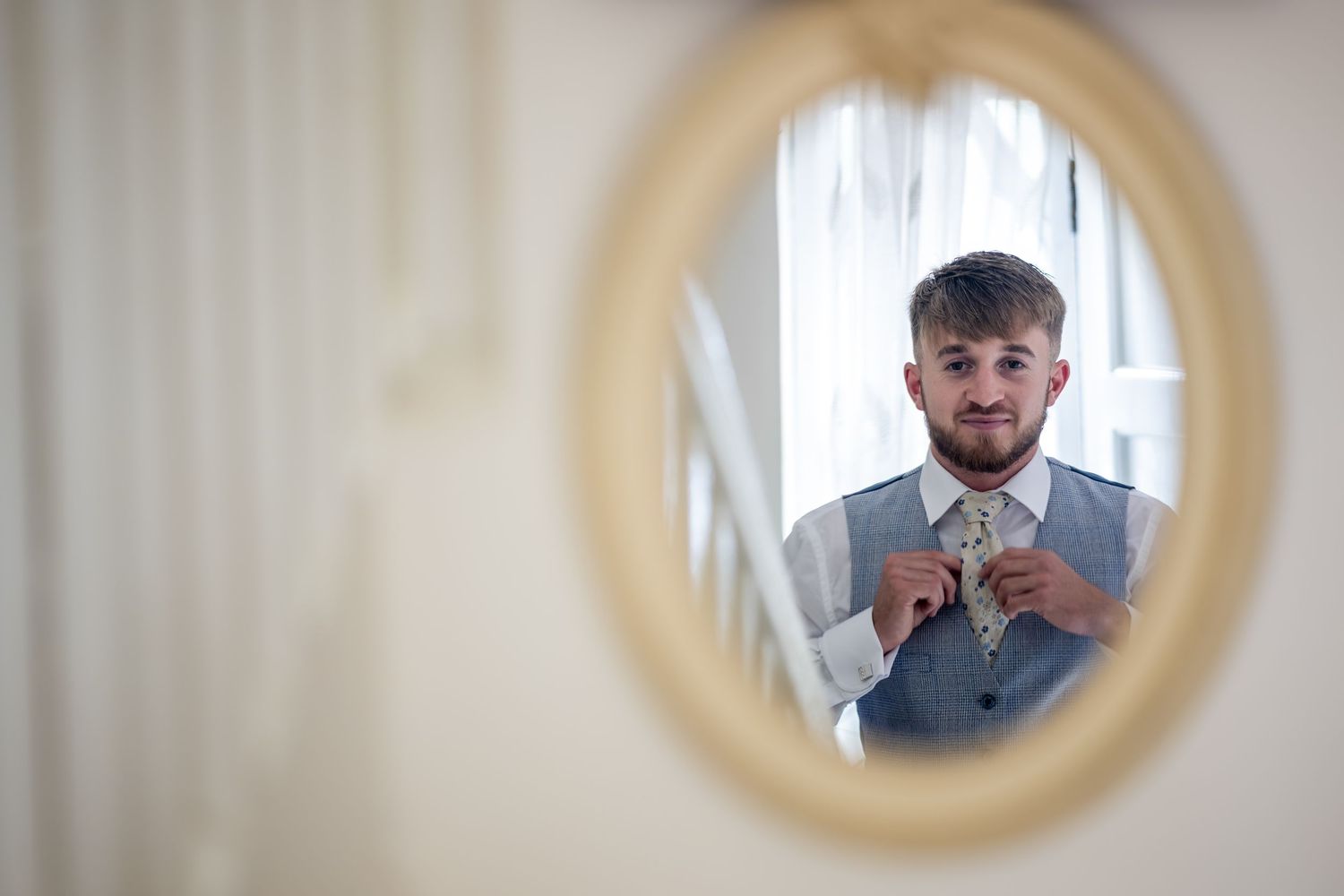 Groom adjusting his floral tie in an elegant oval mirror while wearing a grey vest and white shirt.
