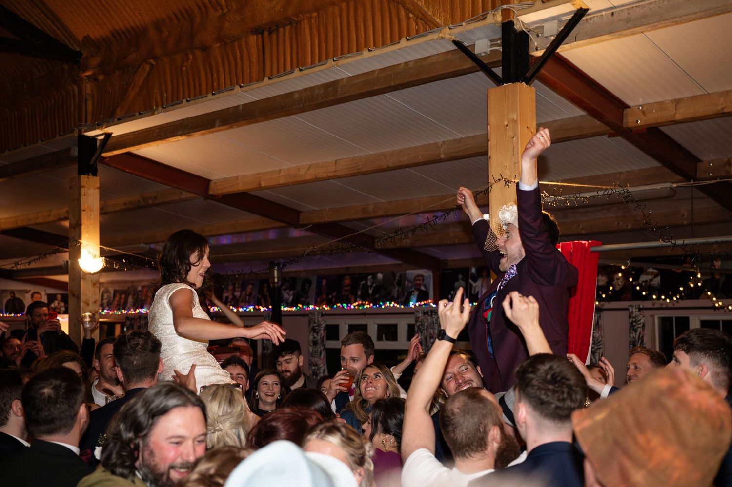 Bride and groom crowd surfing at lively wedding reception party with cheering guests in rustic barn venue.