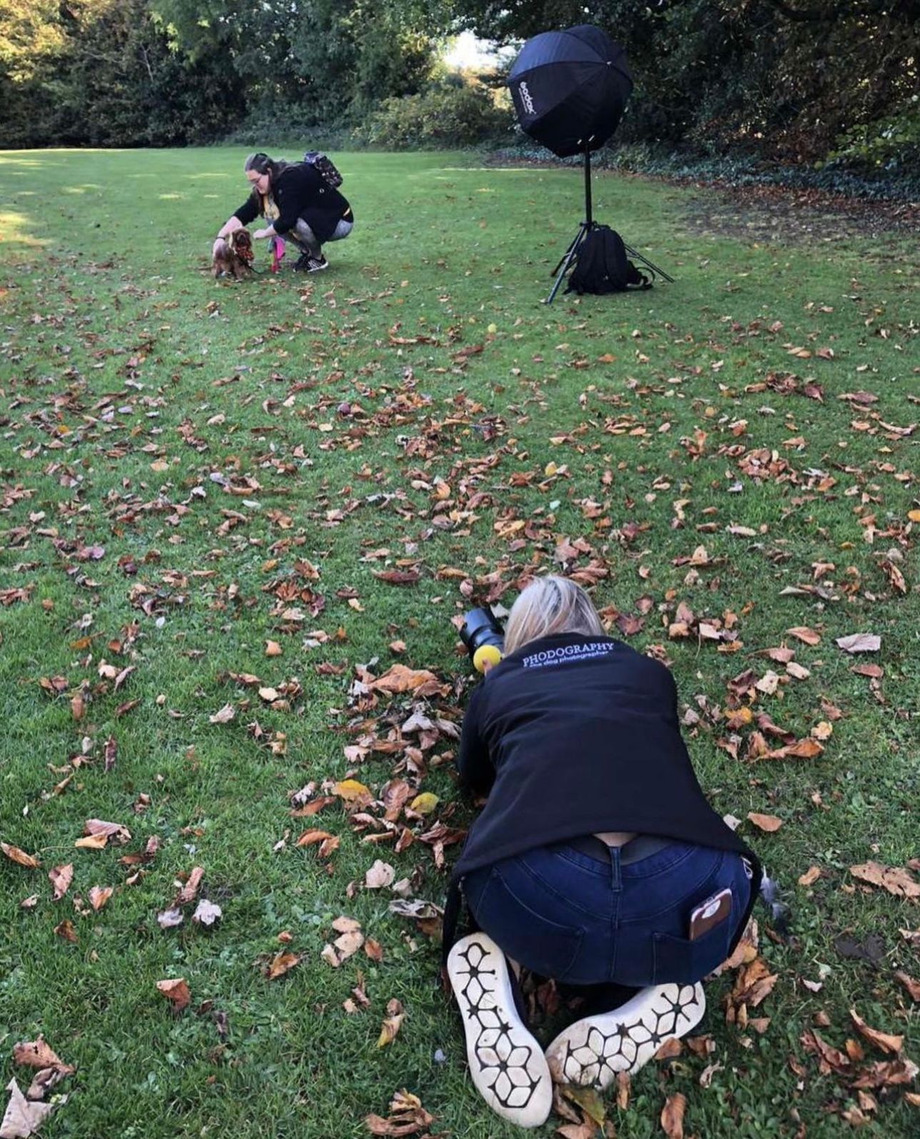 Two photographers lying and crouching on autumn leaves in a garden with studio lighting equipment.