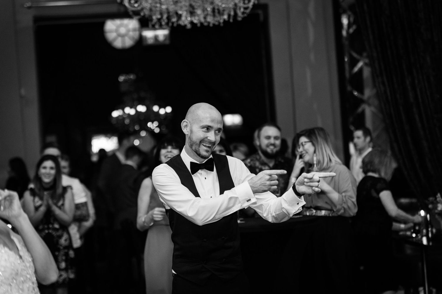 Bald man in vest and bow tie smiling while dancing at elegant black and white wedding reception.