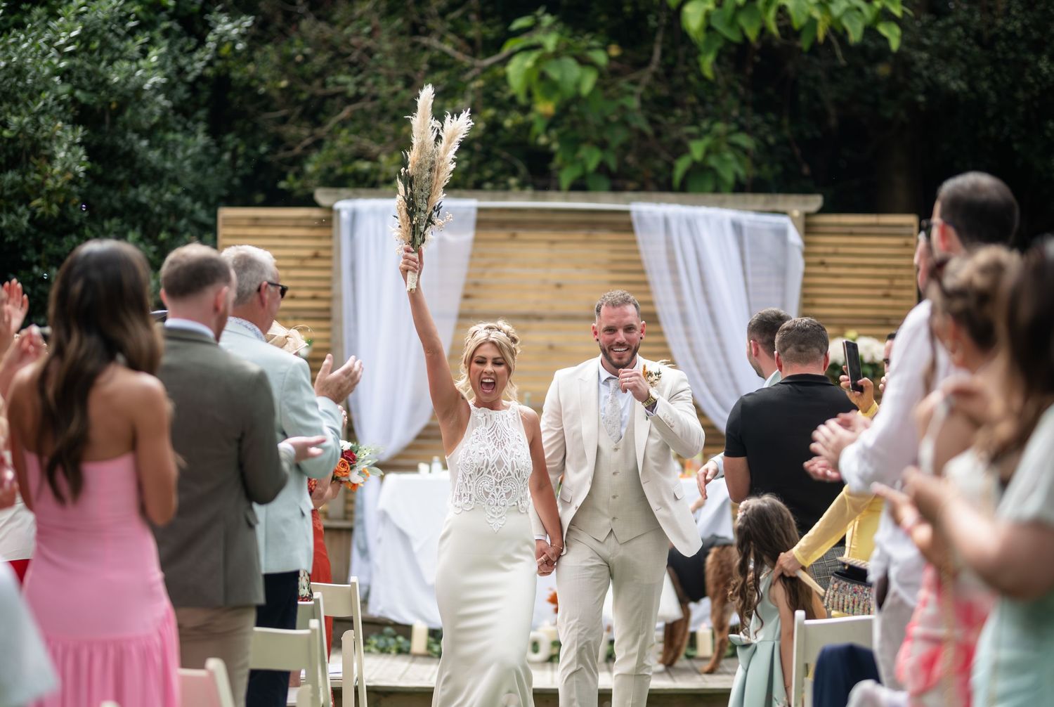 Joyful newlyweds celebrate recessional holding hands under floral arch with cheering wedding guests.